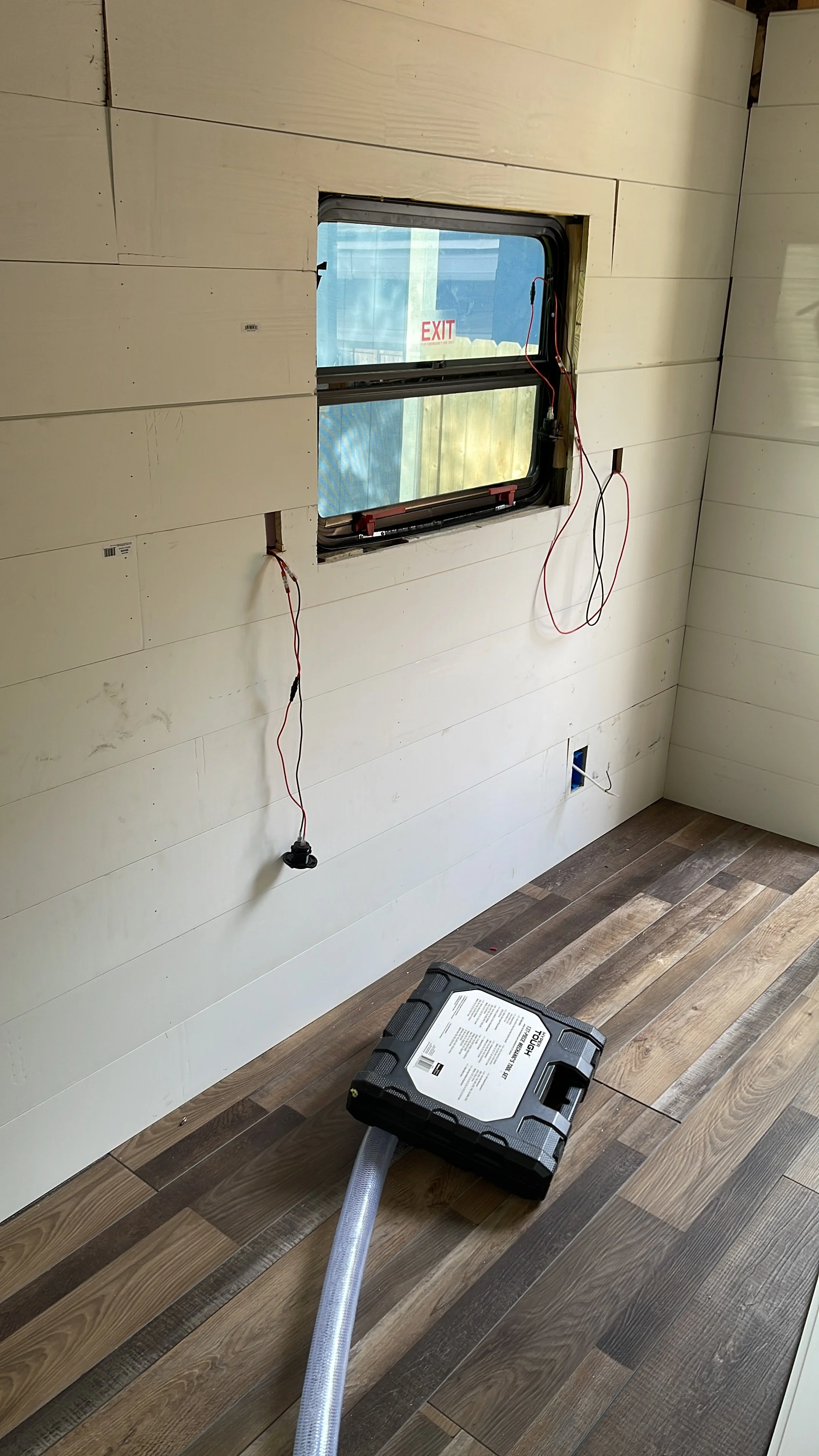 Room under construction with uninstalled window, electrical wires hanging, and a vacuum cleaner on a wooden floor.