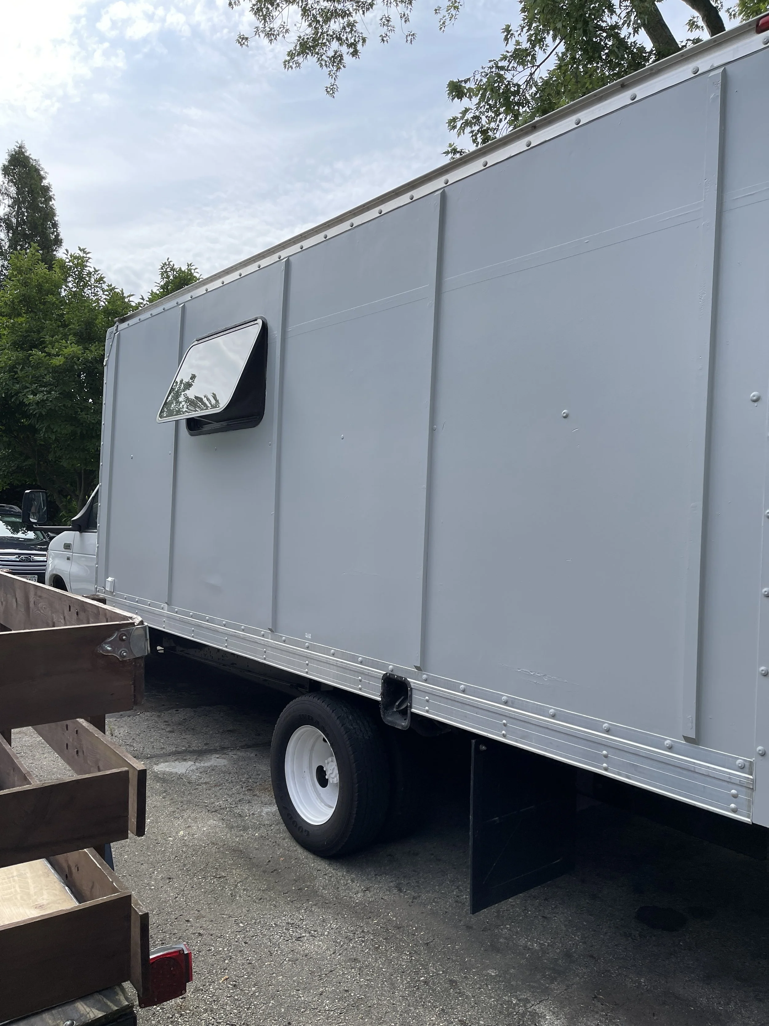 A gray box truck with a side window and black mud flap parked on a gravel surface, with trees and a cloudy sky in the background.