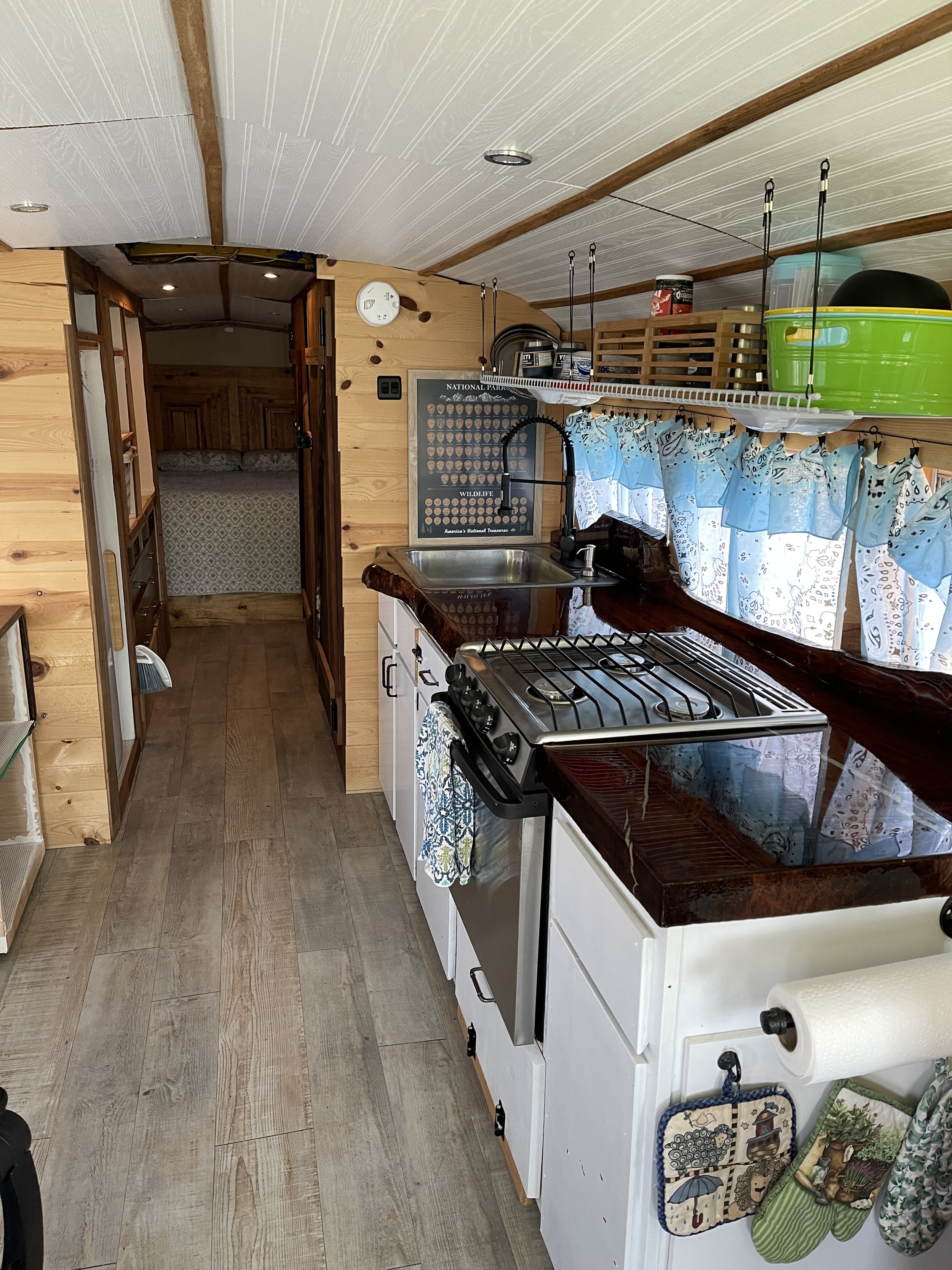 Interior of a small rustic kitchen with wood-paneled walls, a window with blue curtains, a white stove with a dark countertop, and a small sink. There are shelves above the window with various kitchen items and a chalkboard with a wildlife theme on the wall.