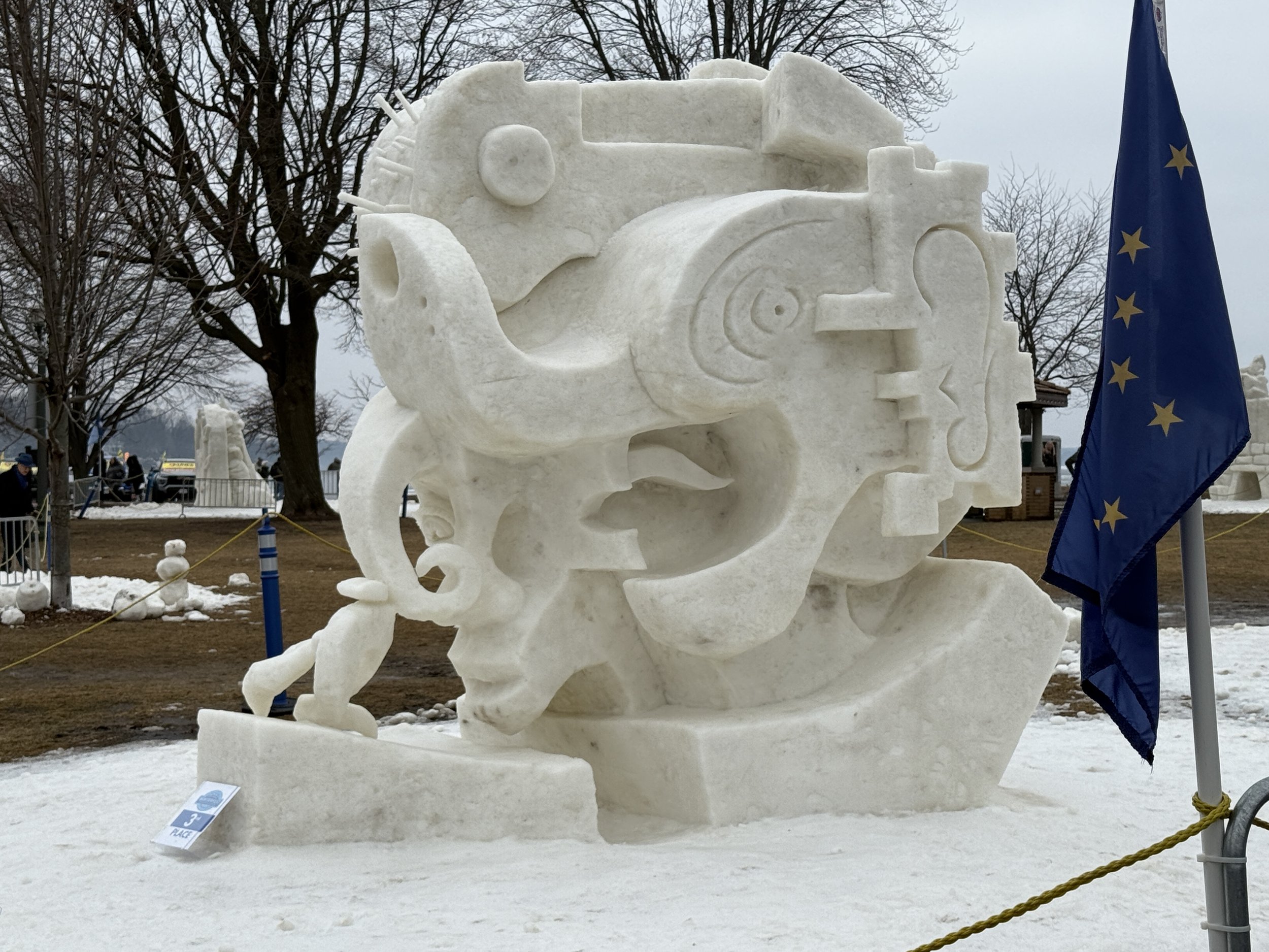 Large snow sculpture resembling a robot face with a European Union flag nearby, outdoors in a park with snow, trees, and a cloudy sky.