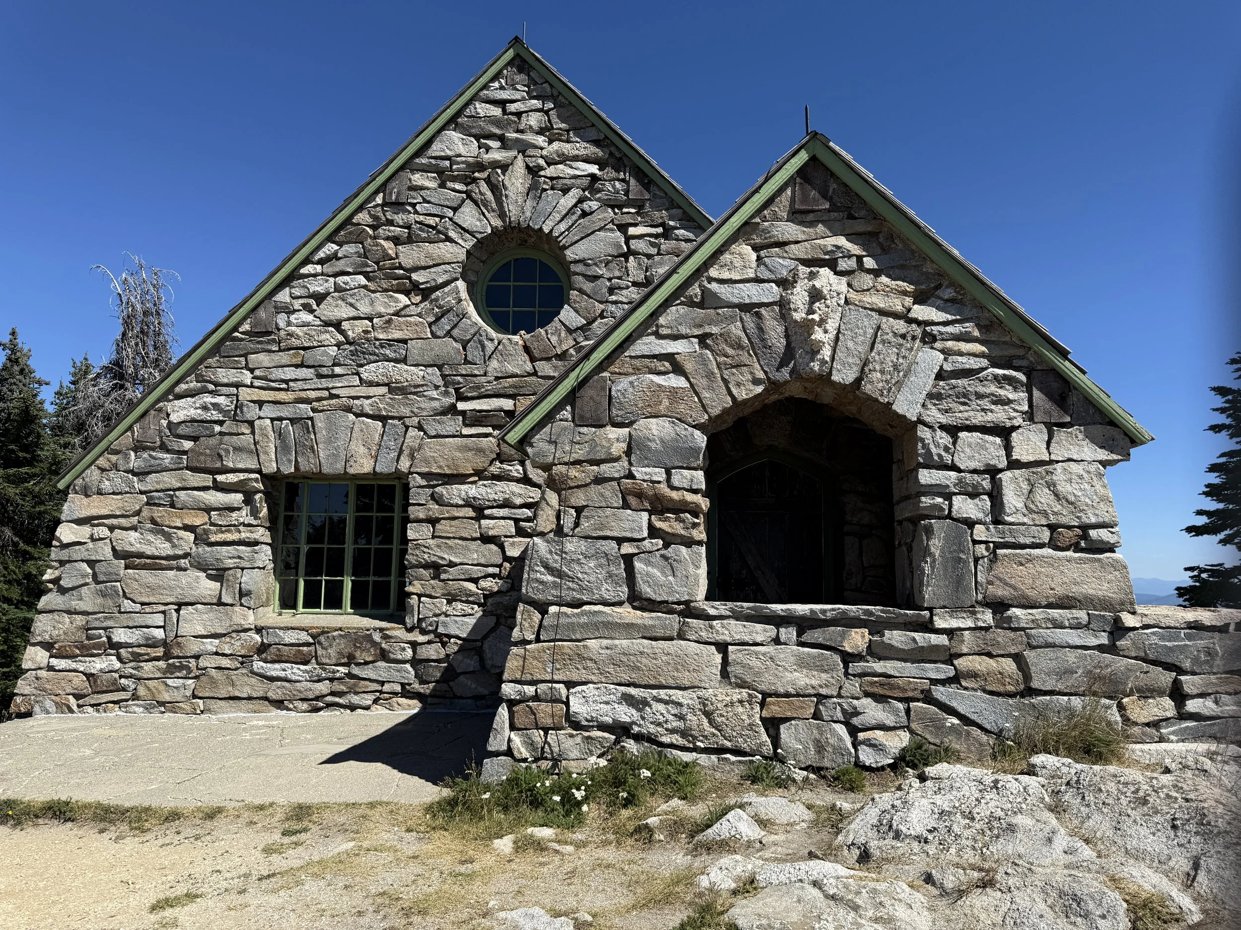 A stone building with green metal framing on the roof, featuring a circular window near the peak, a rectangular window, and an arched entrance, set against a clear blue sky surrounded by trees and rocks.