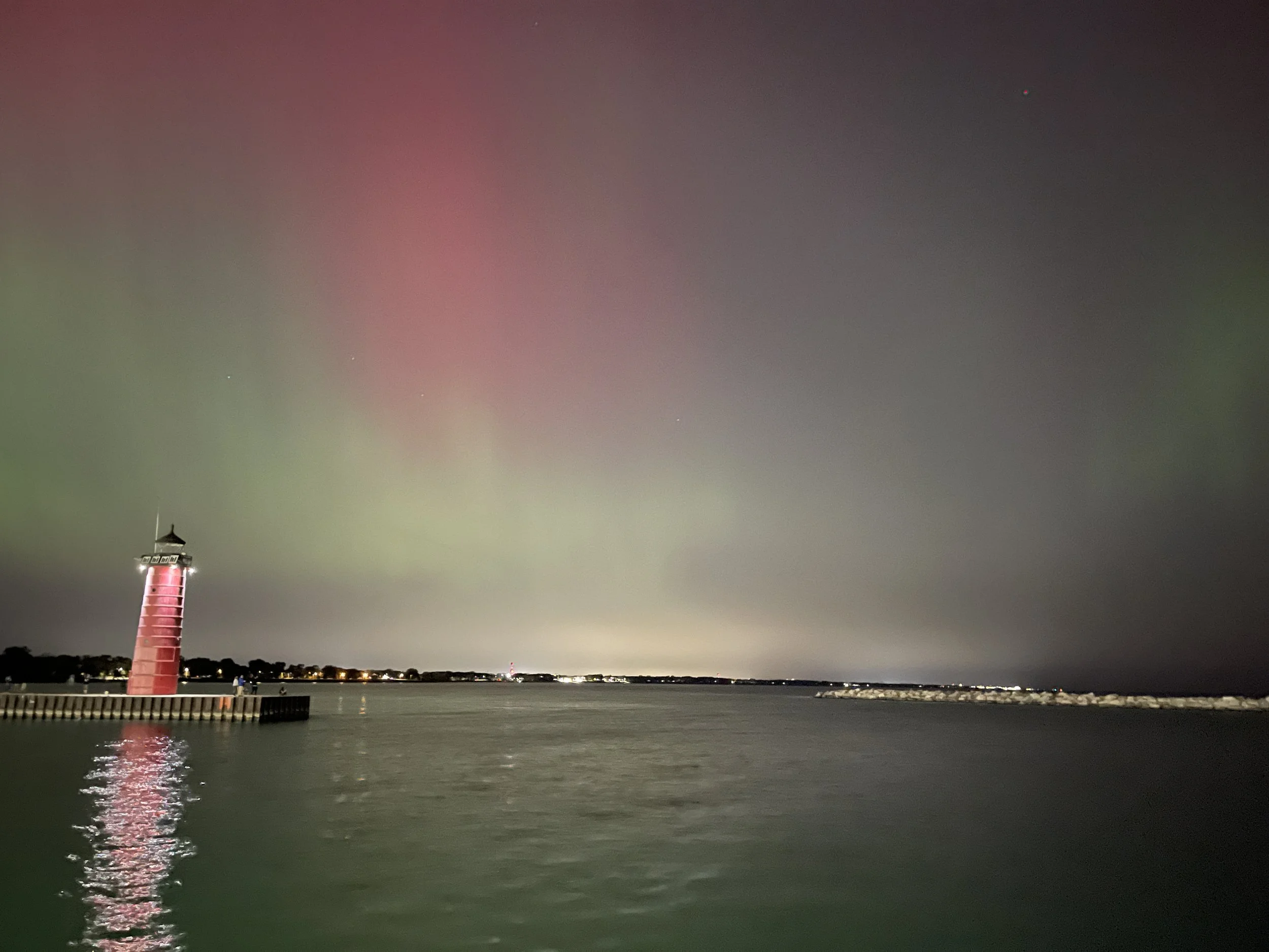 Nighttime scene of a lighthouse on a pier beside water, with a cloudy sky and pink, green, and gray hues, and distant city lights.