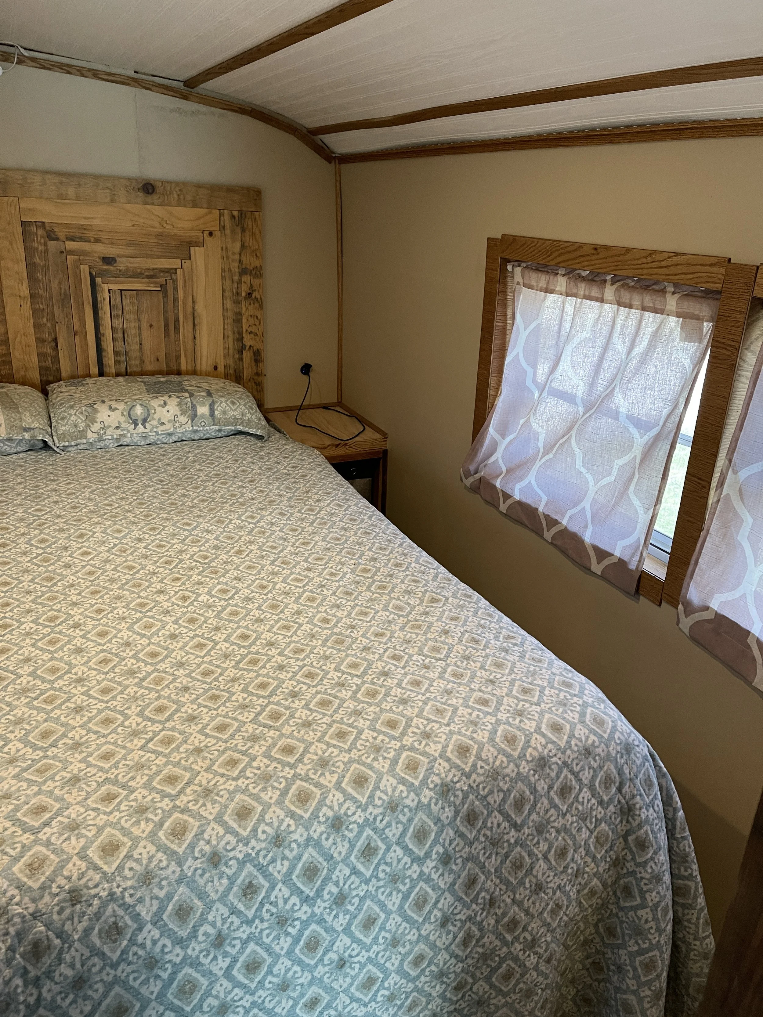 A bedroom with a wooden headboard, patterned curtains, and a window on the right side. The bed has a patterned quilt and pillows.