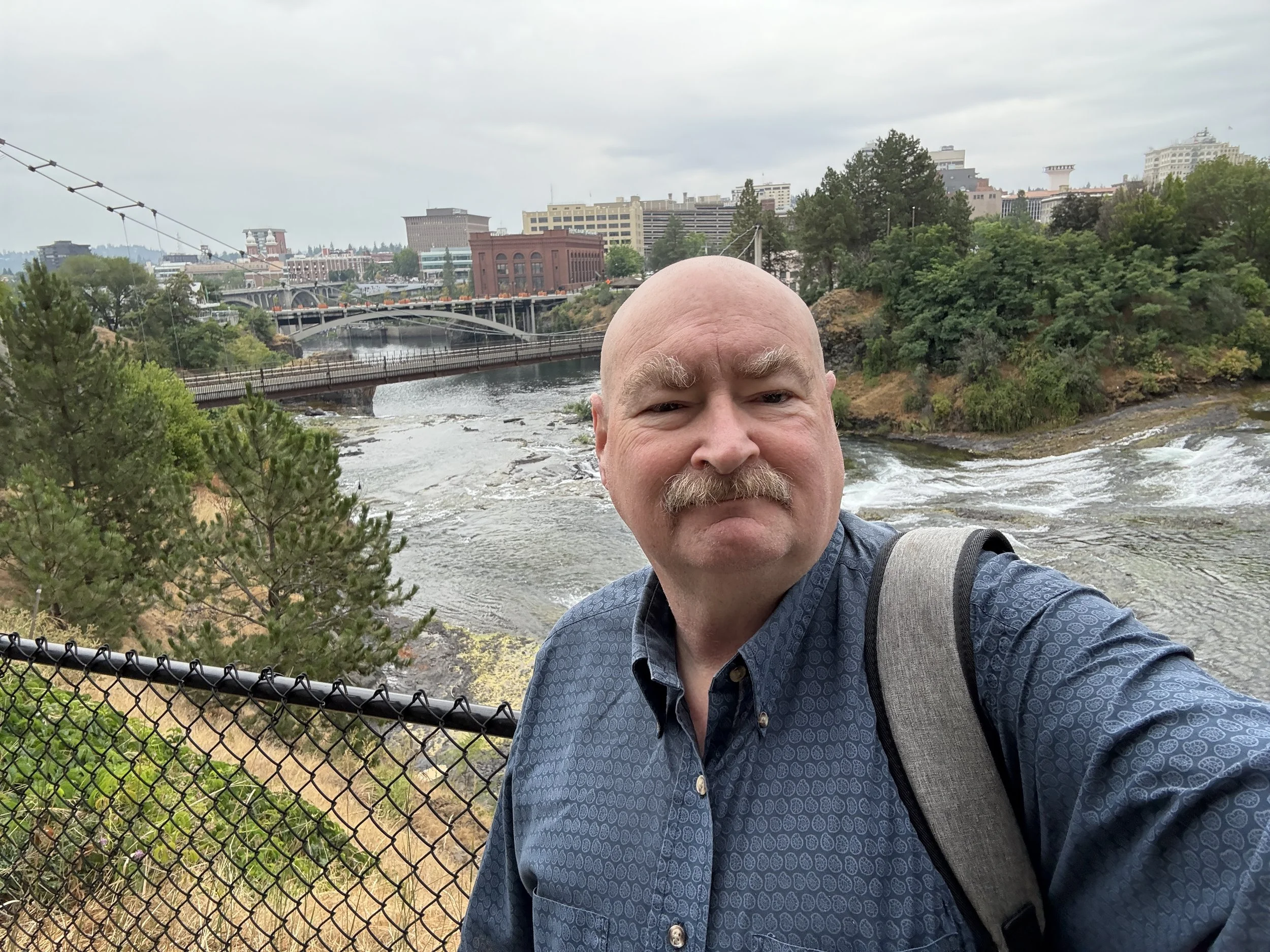 A man with a mustache taking a selfie on a bridge over a river in an urban park, with trees, buildings, and bridges visible in the background.