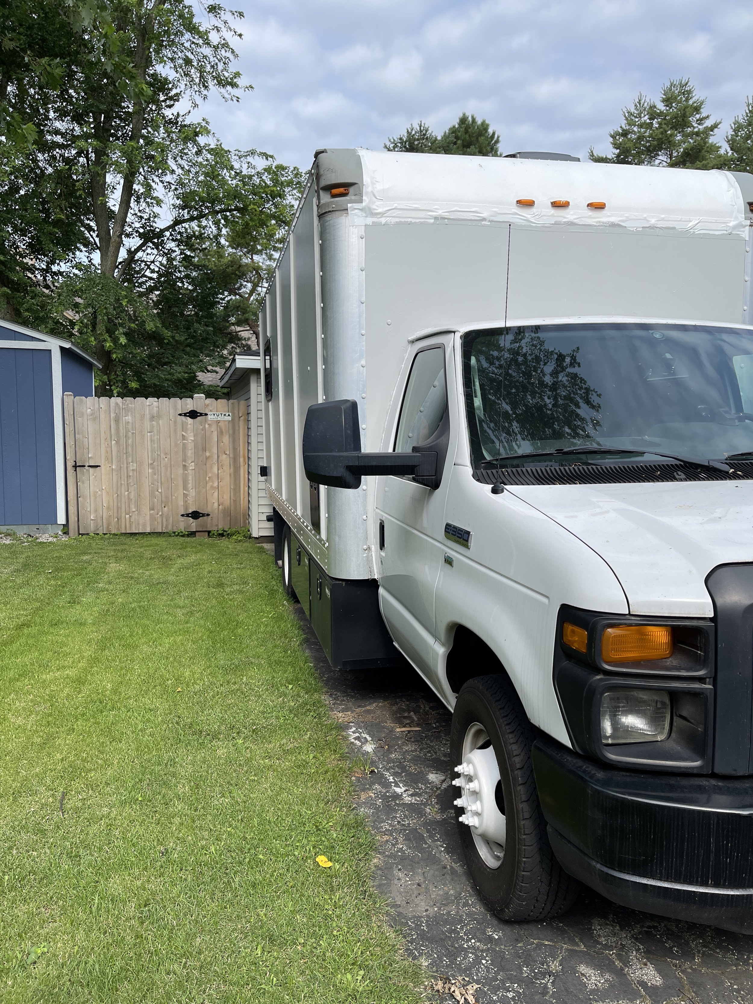 White delivery truck parked in a residential driveway, with trees and a wooden fence in the background.