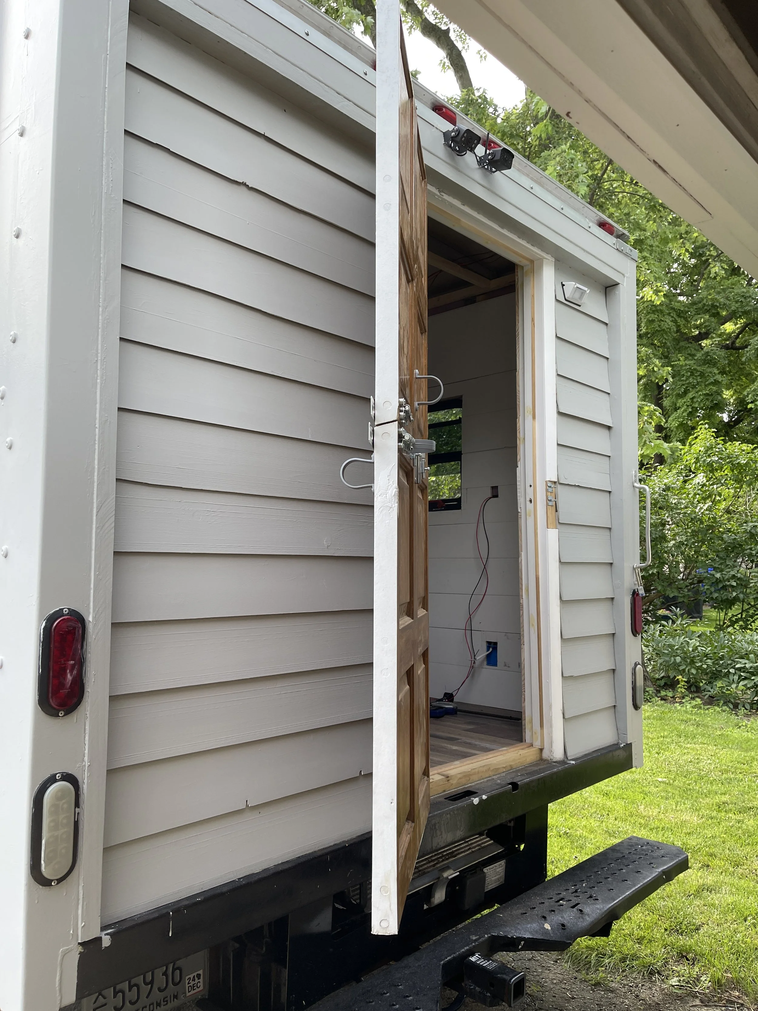 Back of a moving truck with a partially open wooden door, showing an interior space with wiring and an unfinished wall.