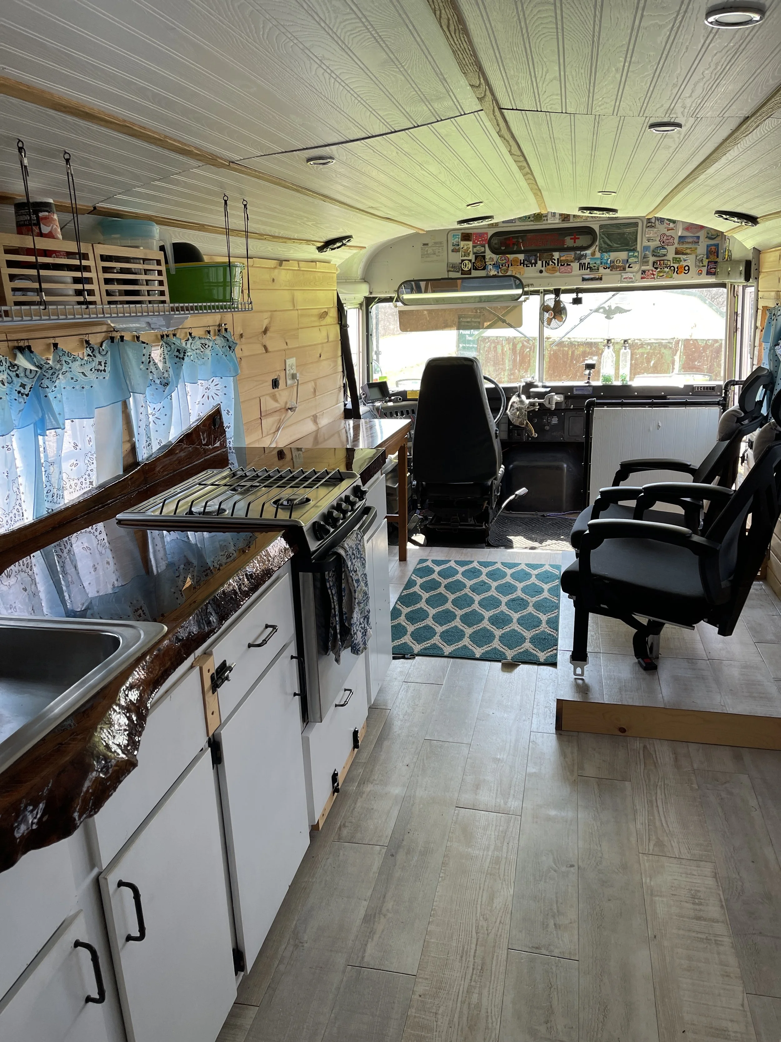 Interior of a converted camper van with a small kitchen on the left, featuring a sink, stovetop, and storage cabinets. The front area has a driver's seat and a passenger seat, with a small table and a blue patterned rug on the floor. The ceiling and walls have wood paneling, and the windshield reveals a view outside with sunlight.