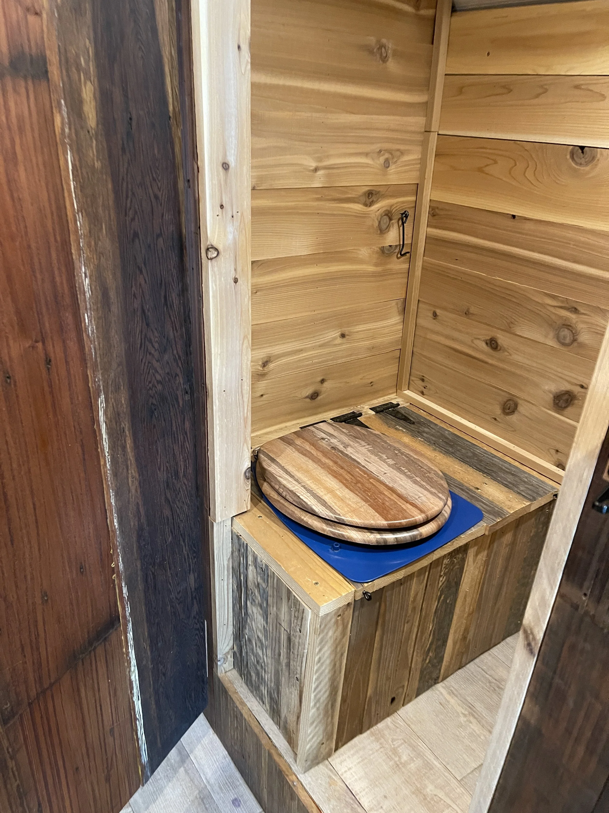 A small wooden cabinet with a hinged door, featuring a rounded wooden seat on top, situated in a wood-paneled room.