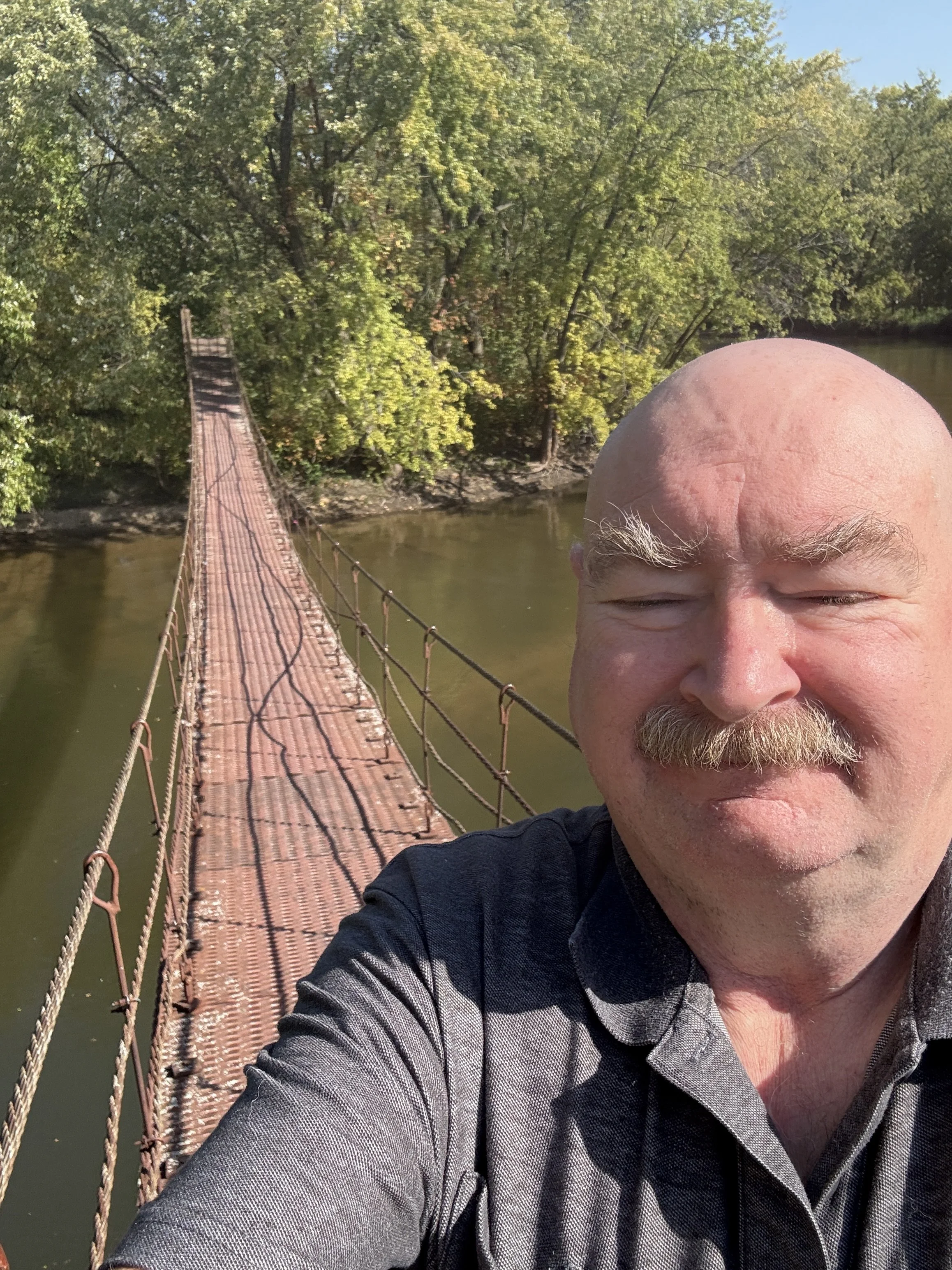 A man with a mustache and bald head taking a selfie on a narrow suspension bridge over a river, surrounded by green trees.