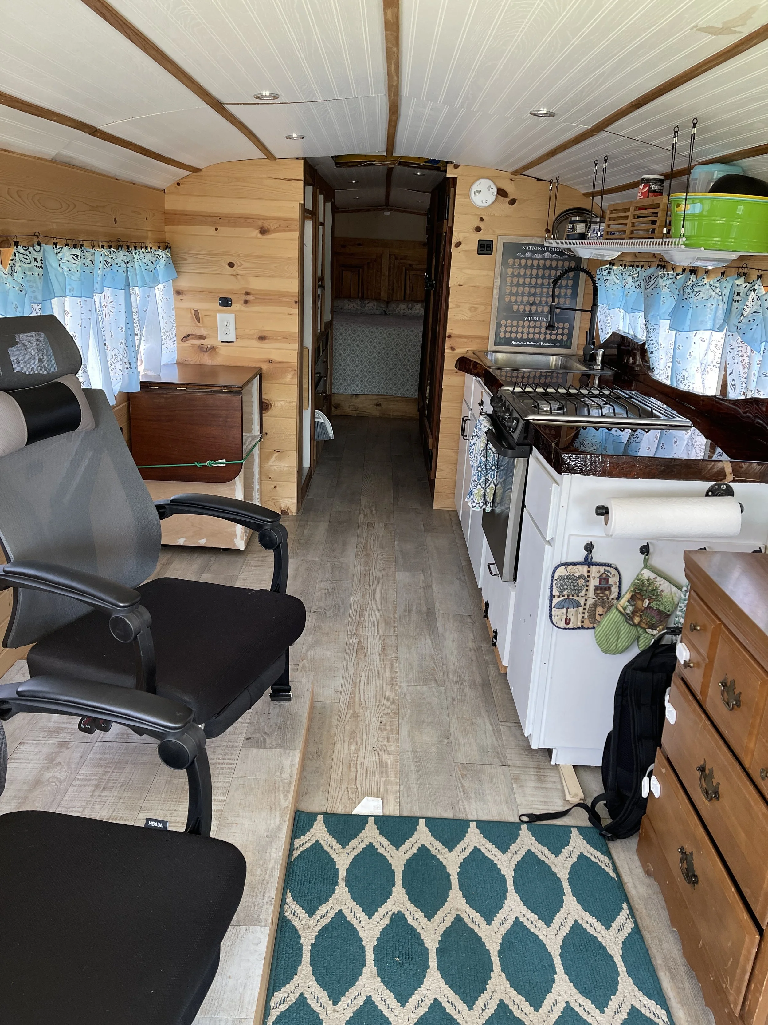Interior of a small wooden camper or tiny house kitchen with a black office chair, wood floors, a rug, a small kitchen counter with a stove and sink, blue patterned curtains, and wood-paneled walls.