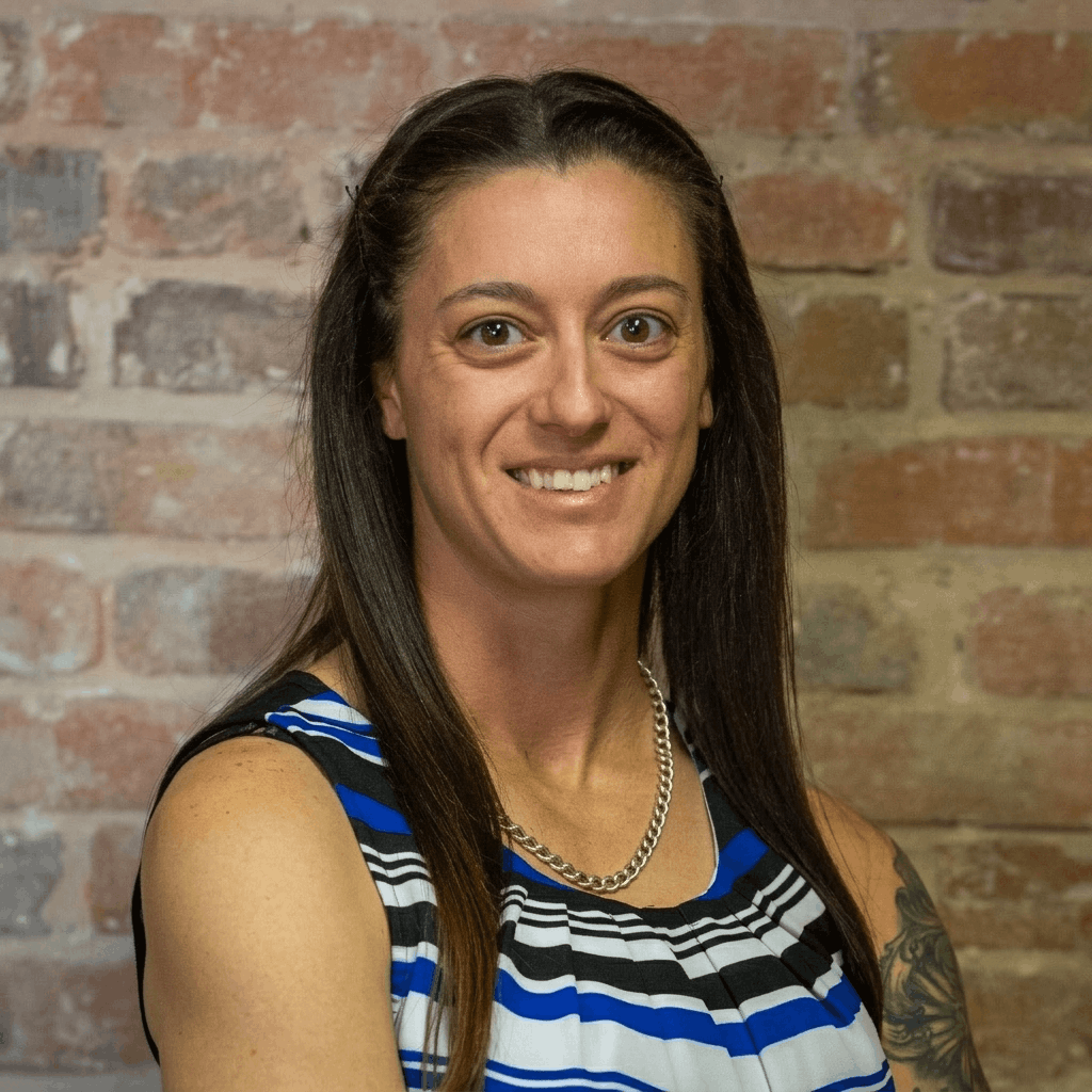 Portrait of a woman with long dark hair, wearing a sleeveless top with blue, black, and white stripes, and a silver necklace, standing against a brick wall background. Shianne Morgan, franchise support.