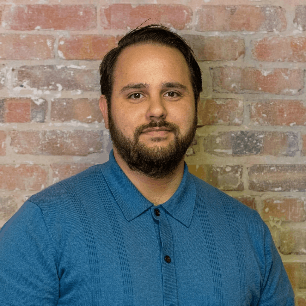 A man with dark brown hair and a beard, wearing a blue collared shirt, standing against a red brick wall. Chris Pilato, franchise support.