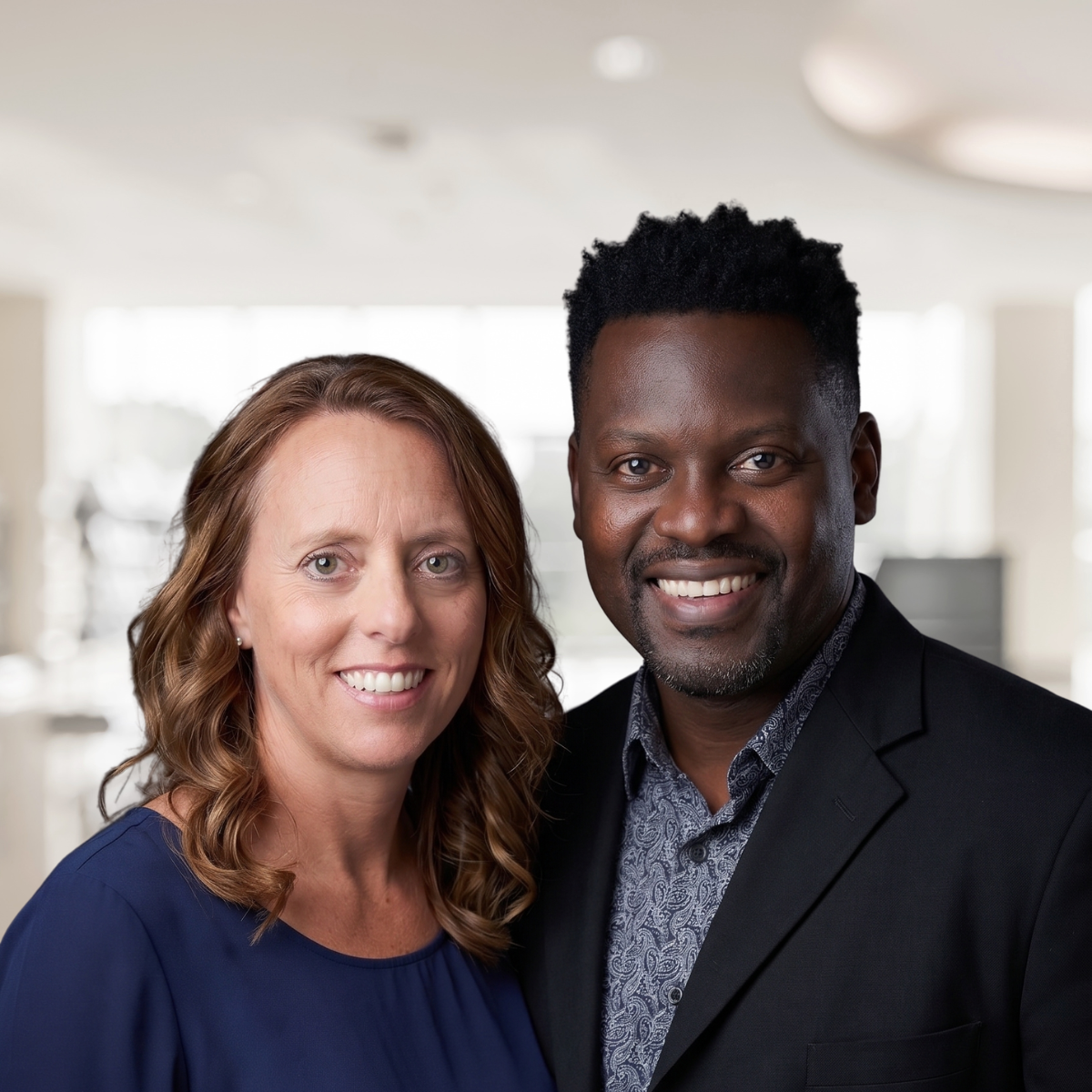 A smiling woman with light skin and wavy red hair wearing a blue blouse, standing beside a smiling man with dark skin and short black hair, wearing a black blazer and patterned shirt, indoors with a blurred bright background. Ed & Christy Ledlum.