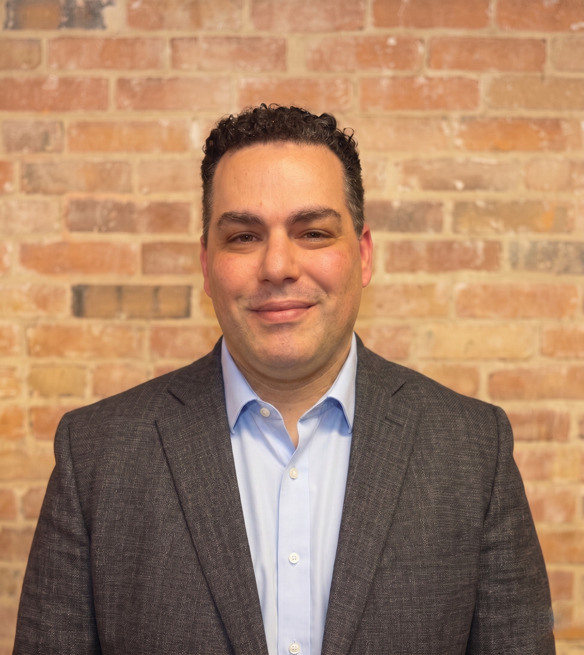 Close-up of a smiling man in a suit against a brick wall. Patrick Capozza, franchise support.