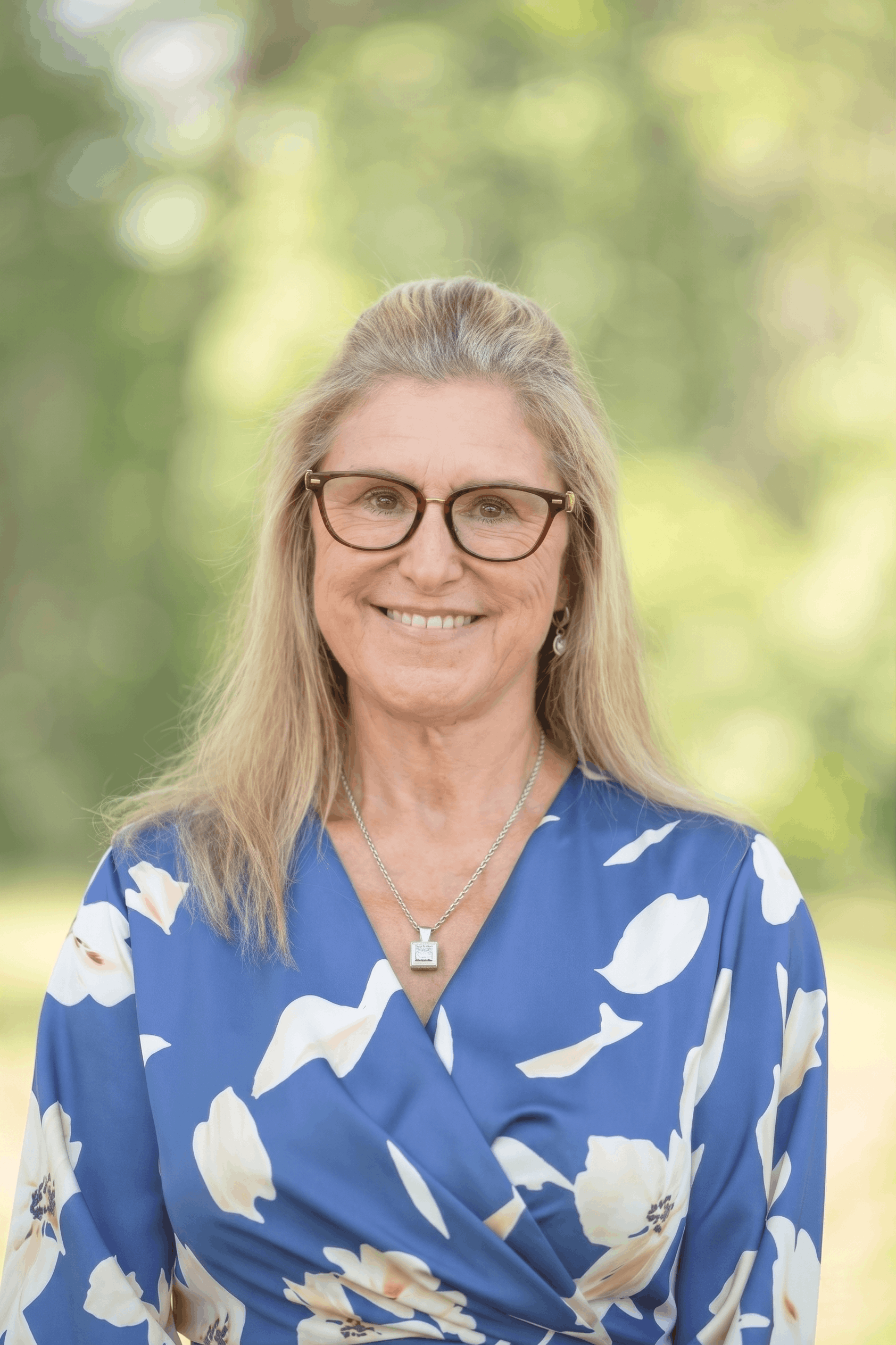A smiling middle-aged woman with long blonde hair, wearing glasses, a blue floral dress, and a necklace, standing outdoors with a blurry green background. Sandy Ferare, co-owner of All County Property.