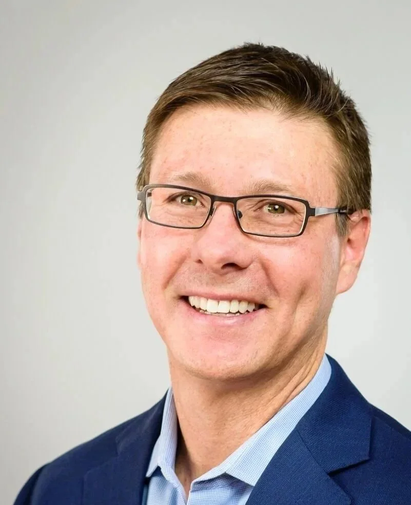 A smiling man with short brown hair, glasses, wearing a blue suit jacket and light blue shirt against a plain light background. Simon Heart, franchise owner in Colorado and Tennessee.