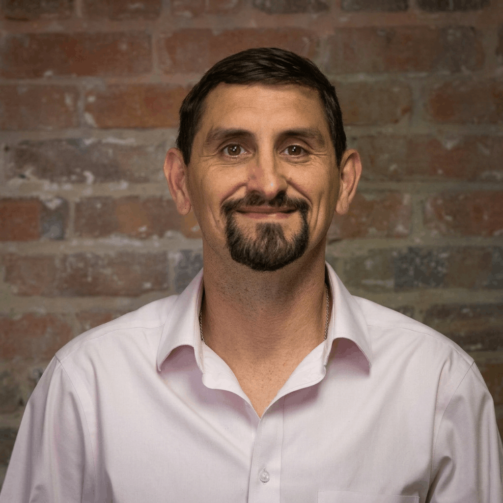 A man with short dark hair, a goatee, and wearing a white collared shirt standing in front of a brick wall. Jerrad McPherson, franchise support.