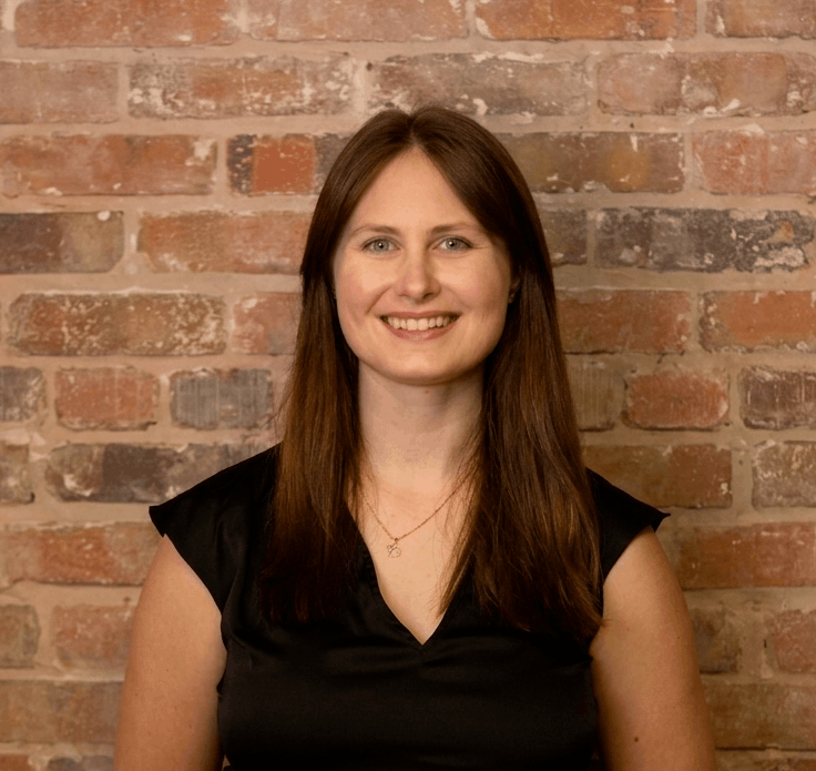 A woman with long brown hair and blue eyes smiling, wearing a black top, standing in front of a brick wall. Sarah McPherson, franchise support.