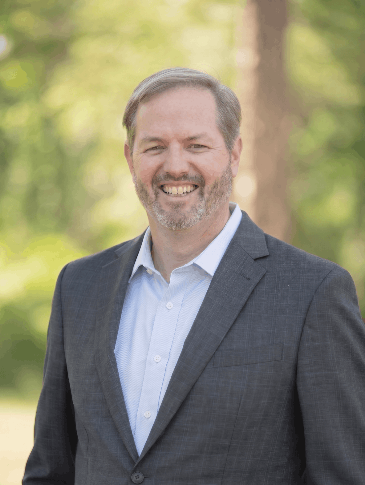 A smiling man with a beard and mustache wearing a dark gray plaid suit jacket and a white button-up shirt, standing outdoors with blurred trees and greenery in the background. Scott McPherson, co-owner of All County Property.