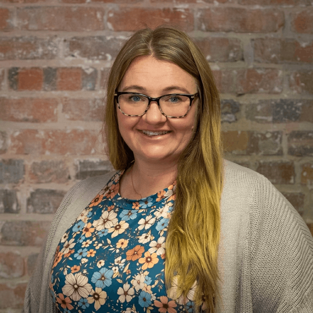 A woman with long blonde hair, glasses, and a floral dress, smiling in front of a brick wall. Lindsey North, franchise support.
