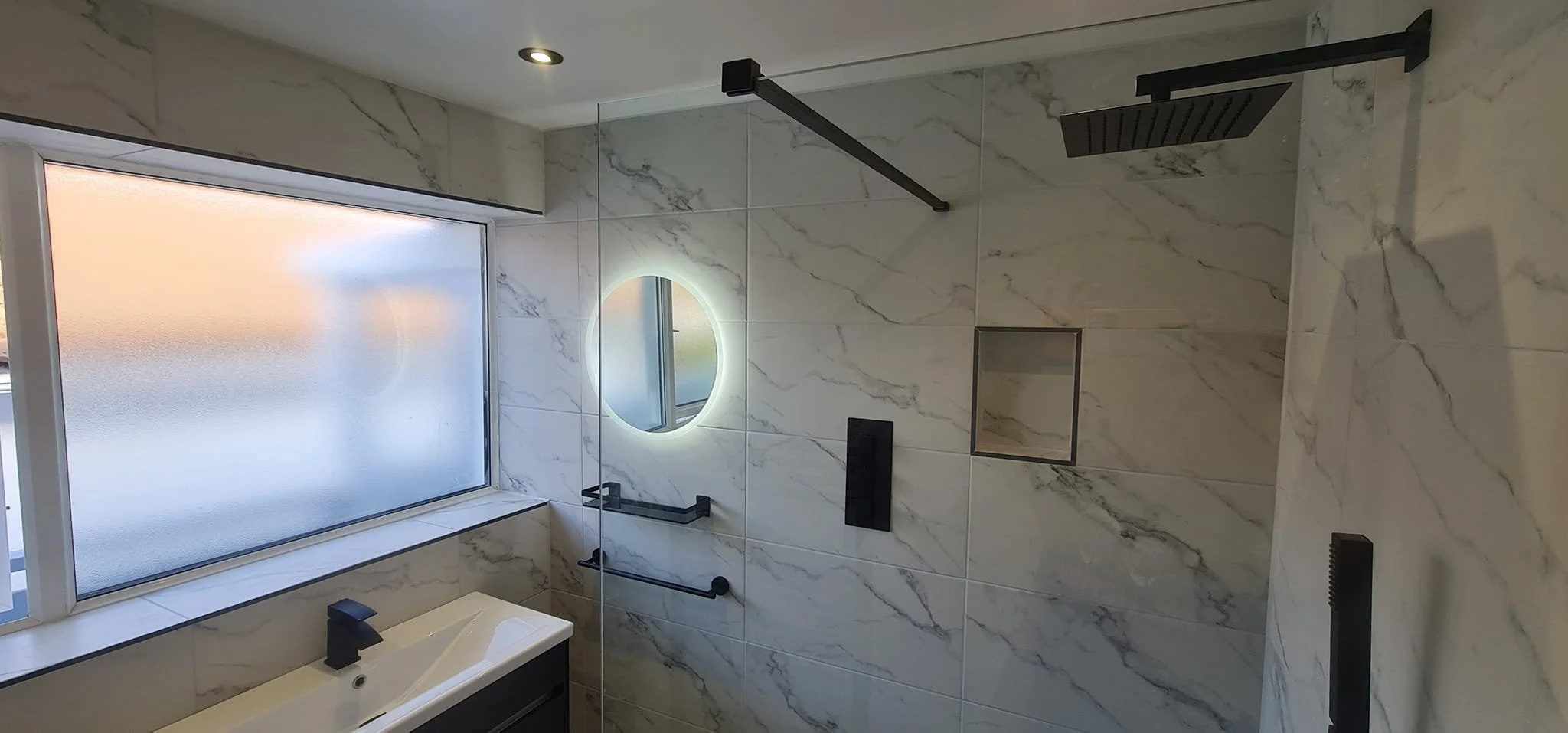 Modern bathroom with marble walls, a walk-in shower with black fixtures, a large frosted window, and a circular backlit mirror above the sink.