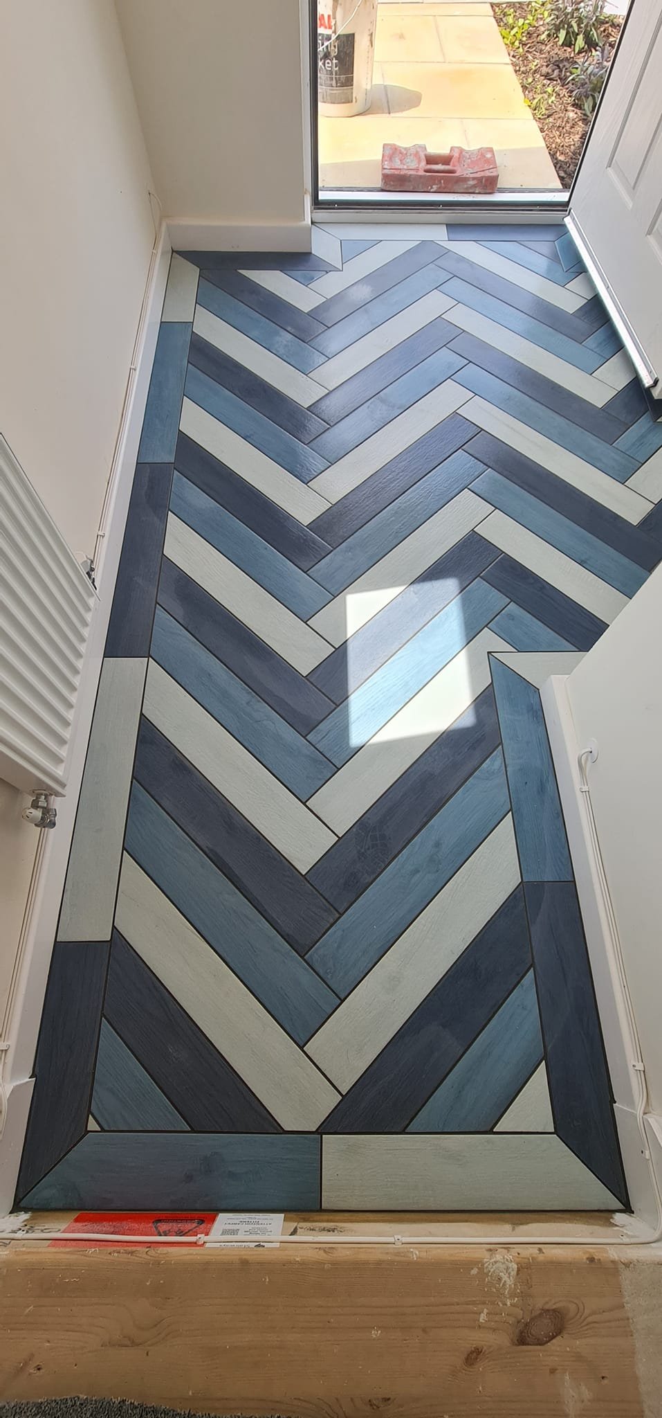 Interior view of a tiled floor with a herringbone pattern in shades of blue and white, near a glass door leading outside to a paved patio area.