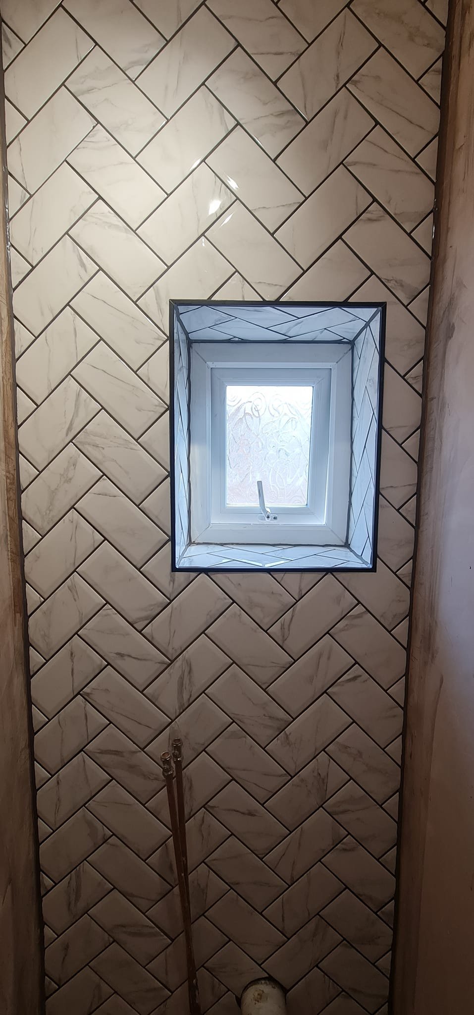 View looking up at a small square window set in a wall with a herringbone pattern of white marble tiles with gray veining. The window has frosted glass and a handle, with black trim around the opening.