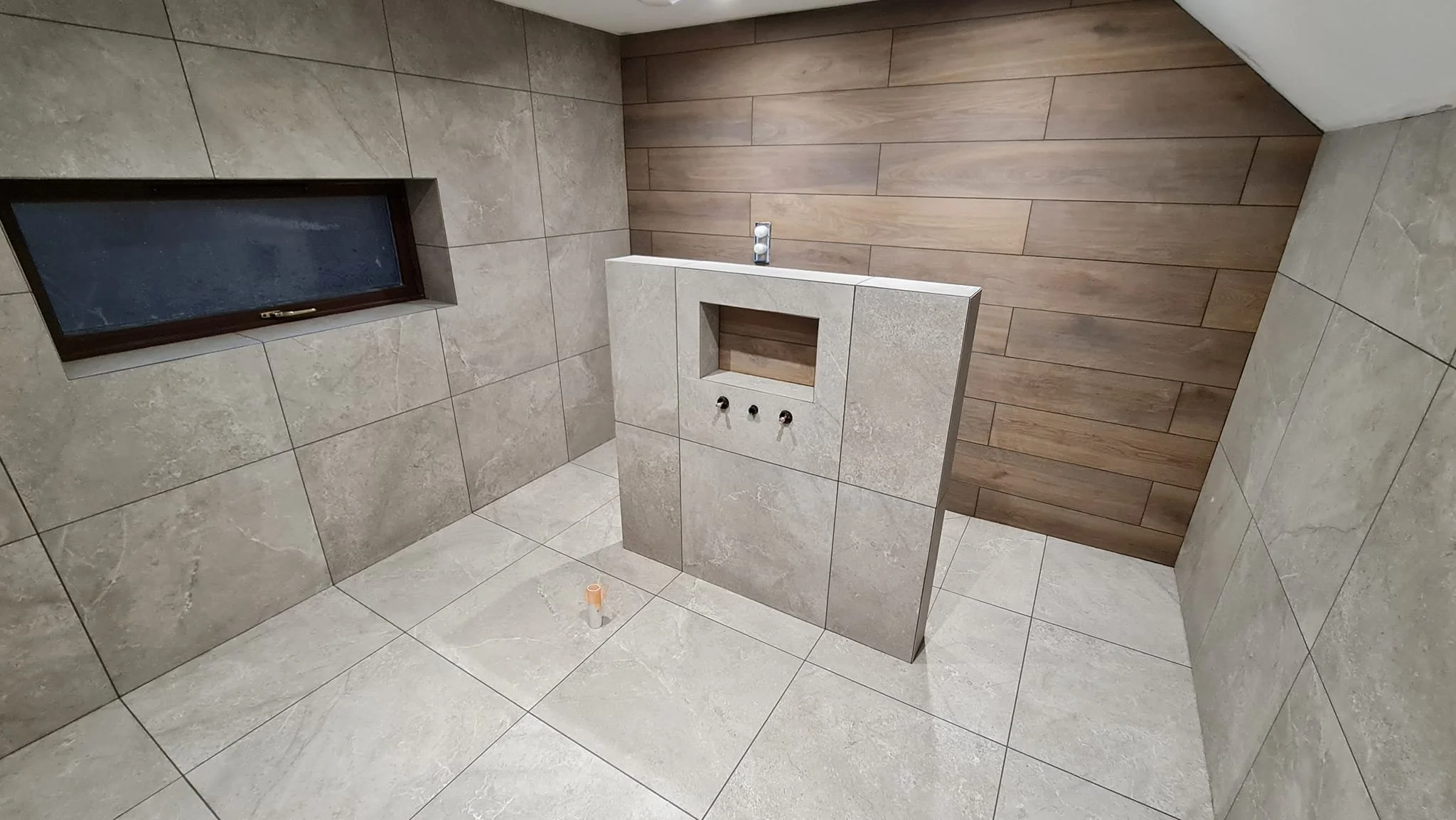 Modern bathroom with beige tiled walls and floor, wooden accent wall, small window, and construction fixtures for a vanity or sink installation.