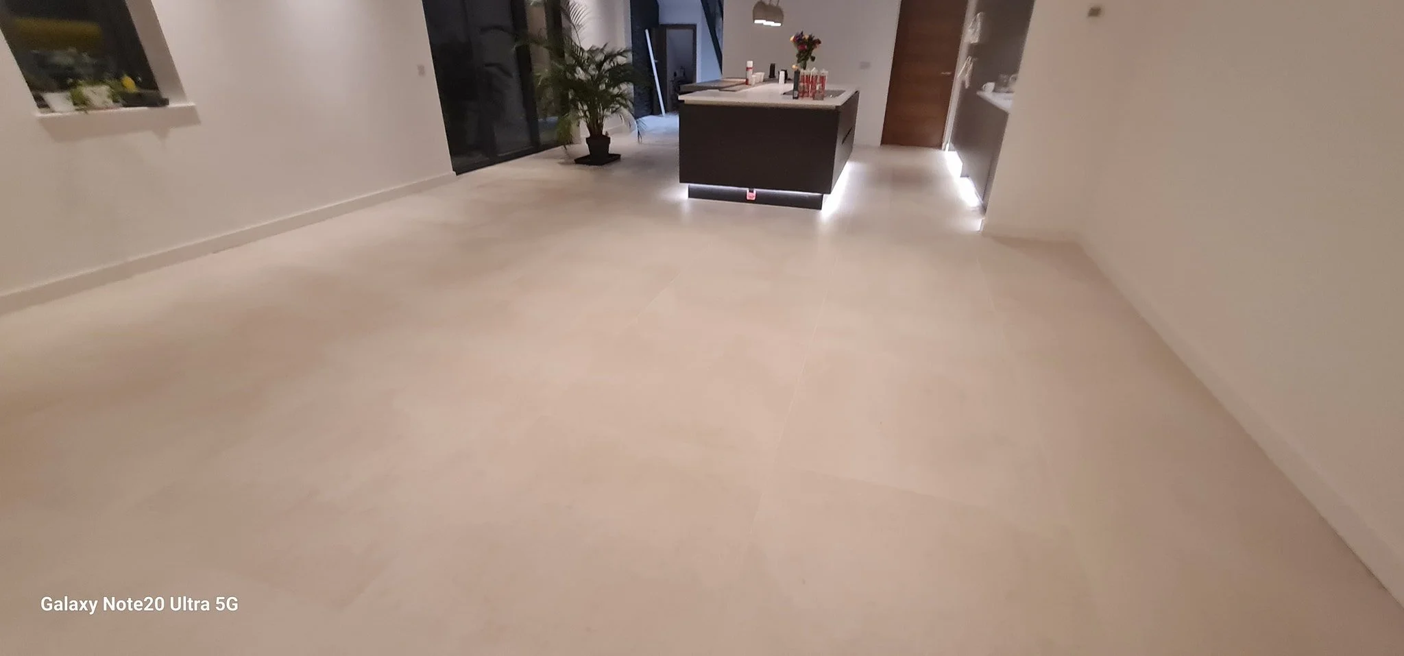Empty room with beige tile flooring, a kitchen island with white countertop, potted plant, and sliding glass door leading outside.