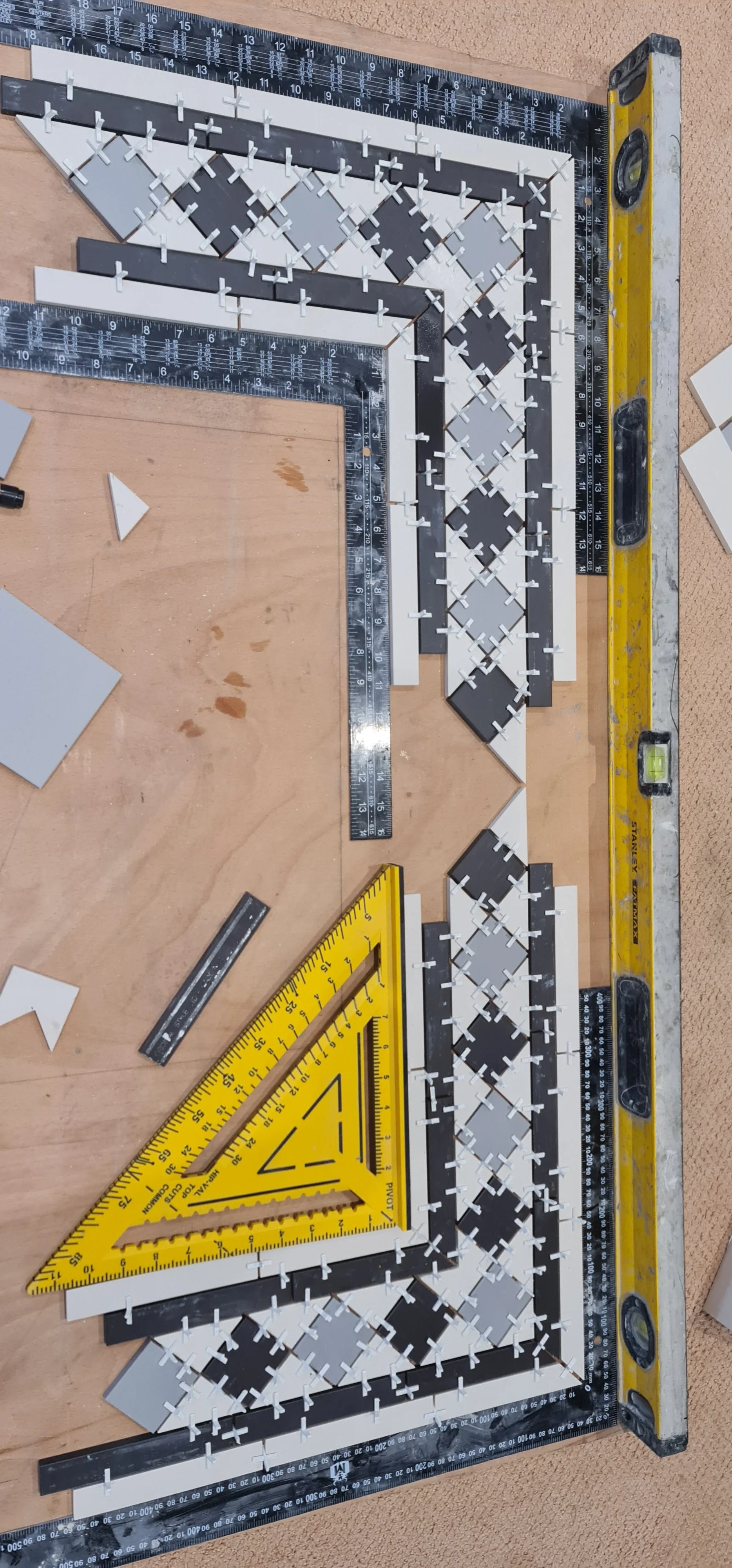 A woodworking setup with a T-square ruler, a yellow triangle ruler, and an array of L-shaped and square tiles arranged in a specific pattern, on a wooden work surface.