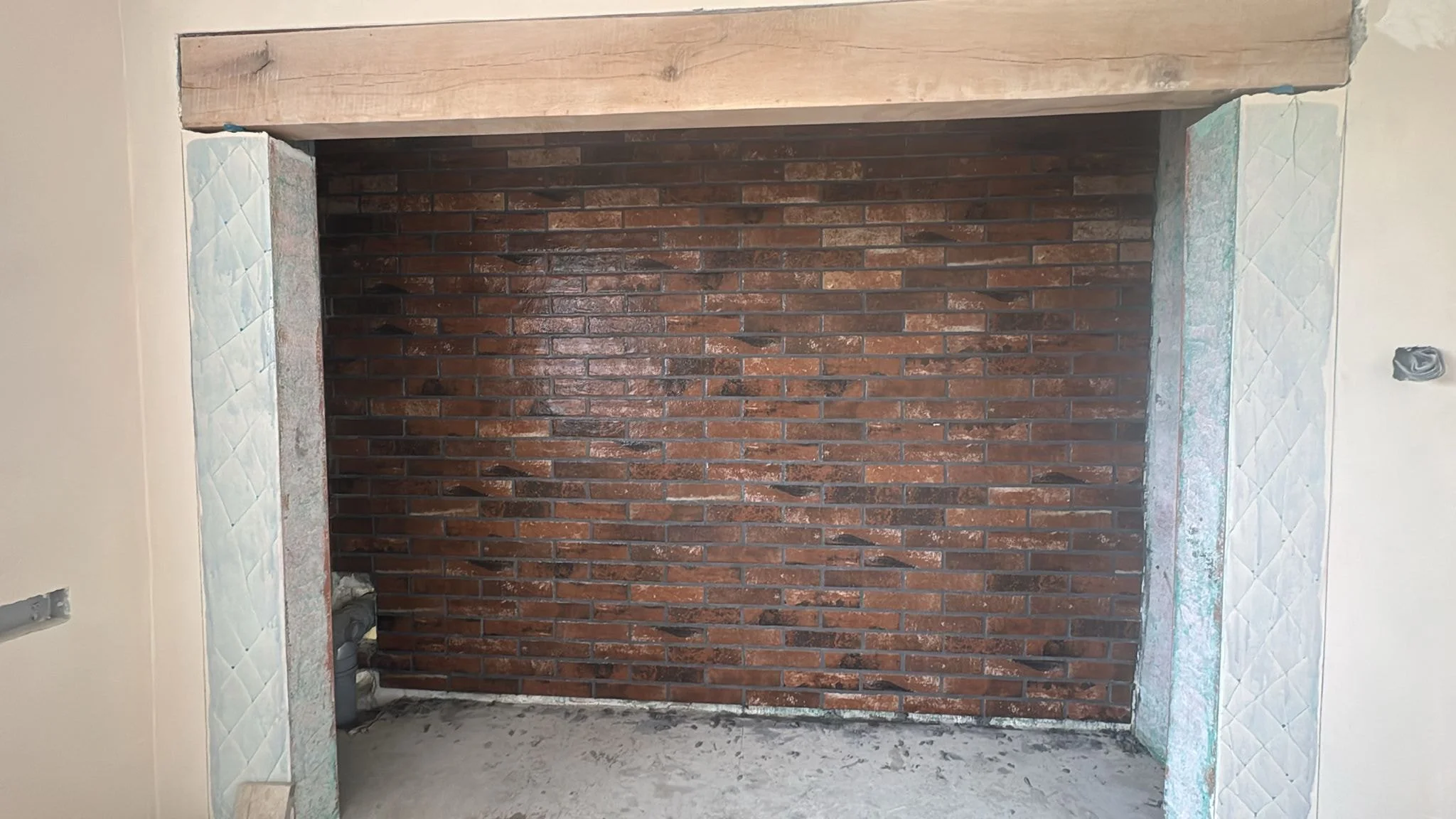 An unfinished room with exposed brick wall and partially installed tiles around the opening, likely for a fireplace or alcove, with visible construction wiring and pipes.