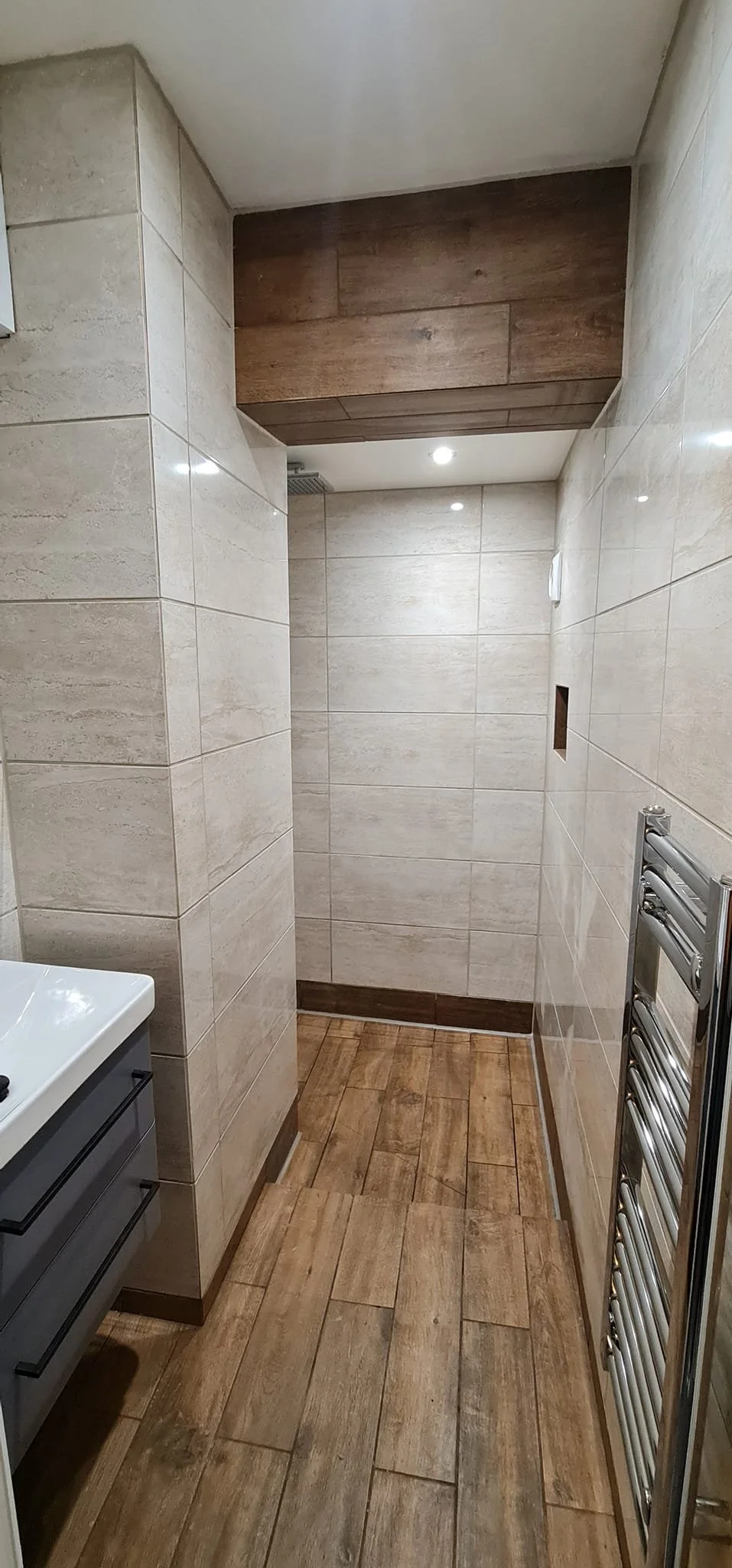 A small, empty bathroom with beige tiled walls, wooden floor, and ceiling, featuring a wall-mounted towel rack, a small sink unit, and a recessed wall niche.