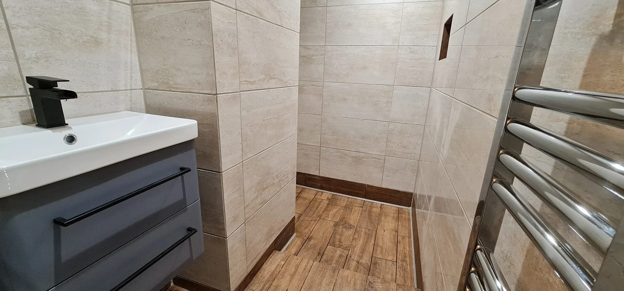 A small bathroom with beige tiled walls and wood-look tile flooring. There's a white sink with a black faucet and storage cabinet below, and a metallic heated towel rack on the right wall.