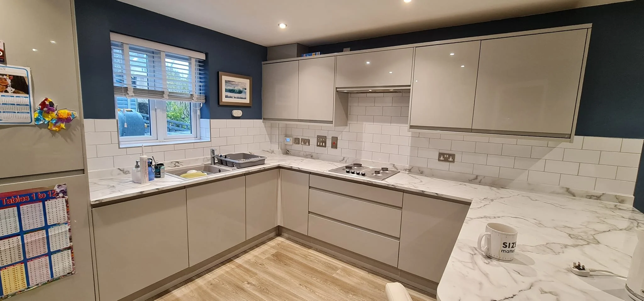 A modern kitchen with white cabinetry and marble countertops. There are small appliances, a dish drying rack, and a coffee mug on the counter. The walls are painted dark blue, and there is a window above the sink with blinds. The backsplash features 