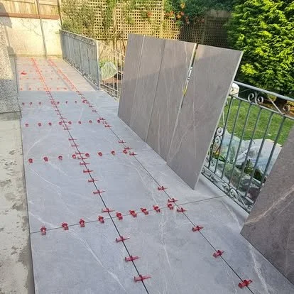 Construction site with tiles and red spacers on a balcony or terrace, with some large tiles leaning against a railing and a green outdoor area in the background.