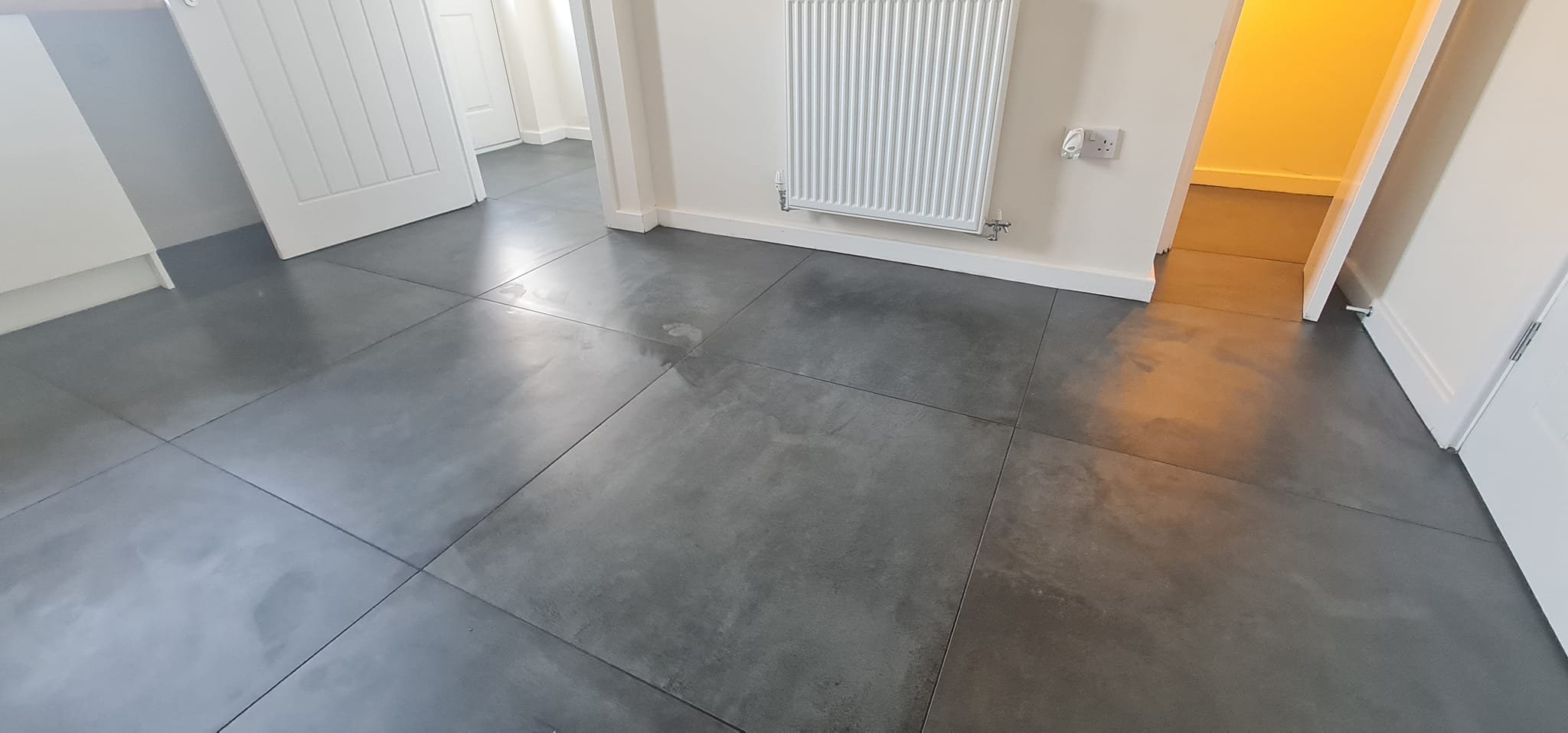 Inside a house showing a tiled gray floor, a white door, a wall-mounted radiator, and a doorway leading to a room with wooden flooring.