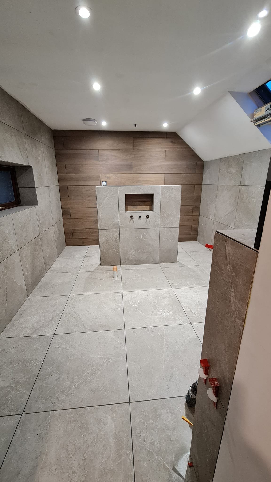 Construction site of a bathroom with incomplete fixtures. The photo shows a tiled floor, tiled walls, a partial wall with plumbing fixtures, and some construction tools and materials.