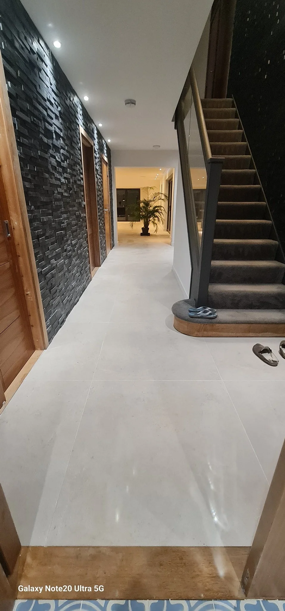 Interior hallway of a modern house with a black textured wall on the left, white tiled flooring, a staircase with brown carpeted steps on the right, and a potted plant in the background near large windows.