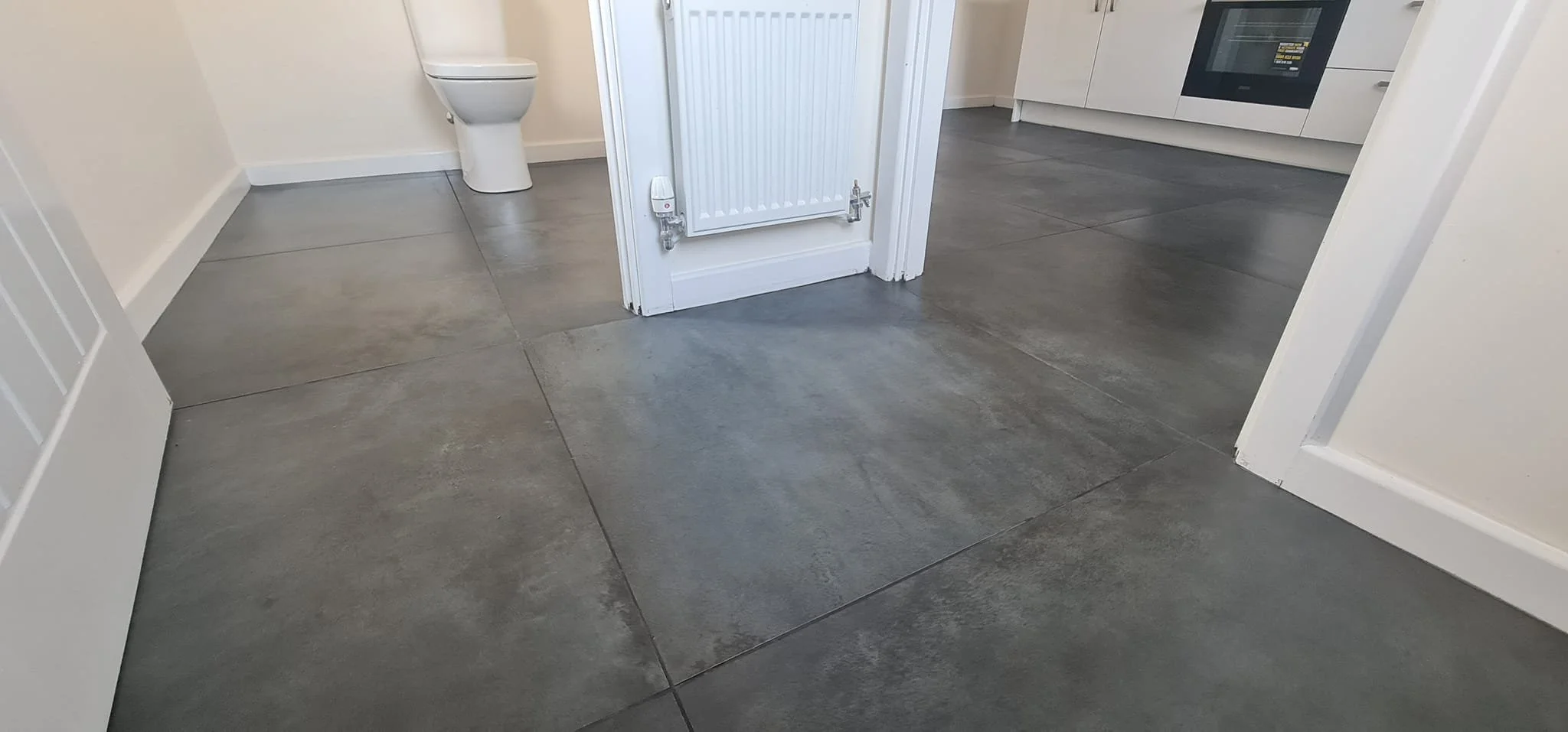 Interior view of a modern living space showing a grey tiled floor, a bathroom with a toilet, and part of a kitchen with white cabinets and a built-in oven.