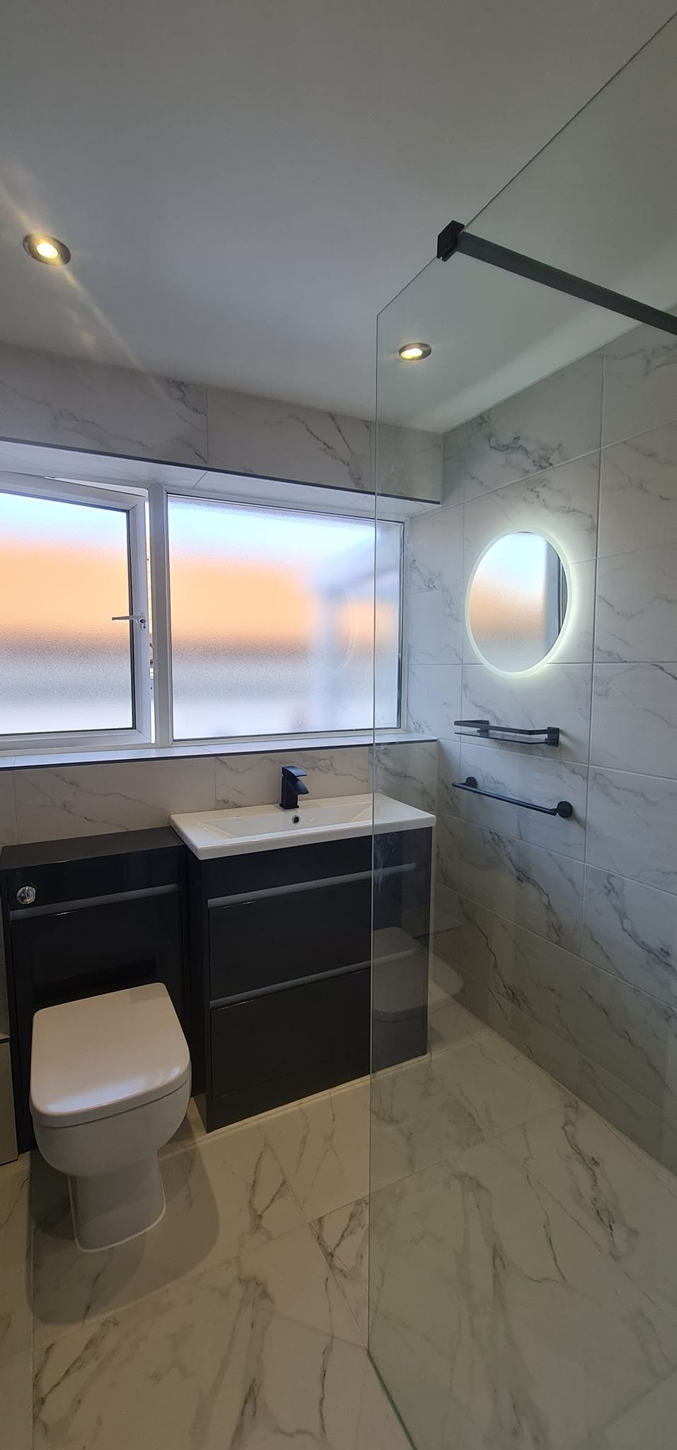 Modern bathroom with marble walls and floor, a black vanity with a white sink, a toilet, frosted window, illuminated round mirror, and towel bars.