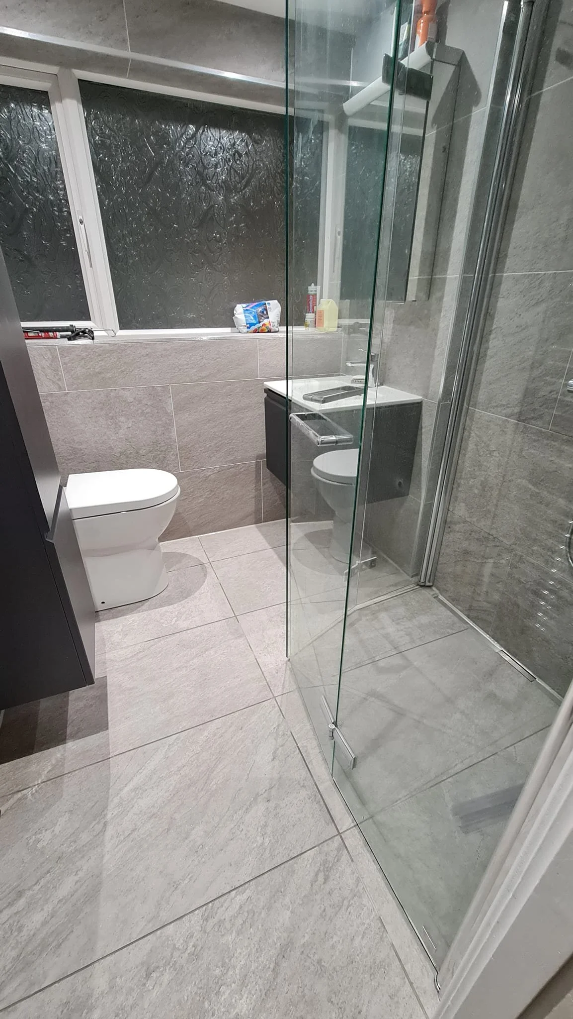 A modern bathroom with gray tiles, a white toilet, a black vanity with a sink, and a glass-enclosed shower stall. The window in the background has frosted glass, with various toiletries and cleaning supplies on the windowsill.