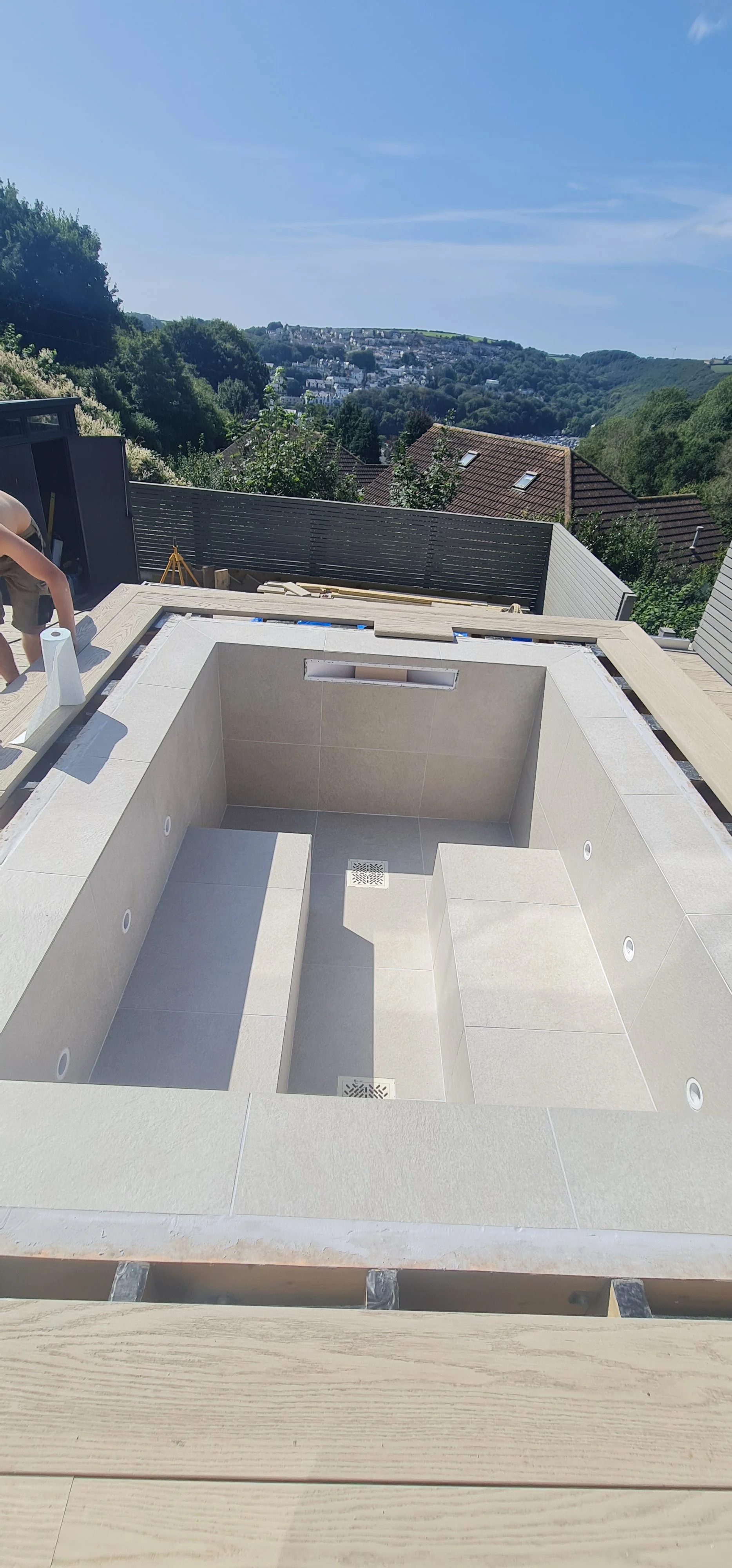 Construction site with an empty hot tub or pool with beige tiles, on a rooftop overlooking a scenic landscape of hills and houses under a clear blue sky.