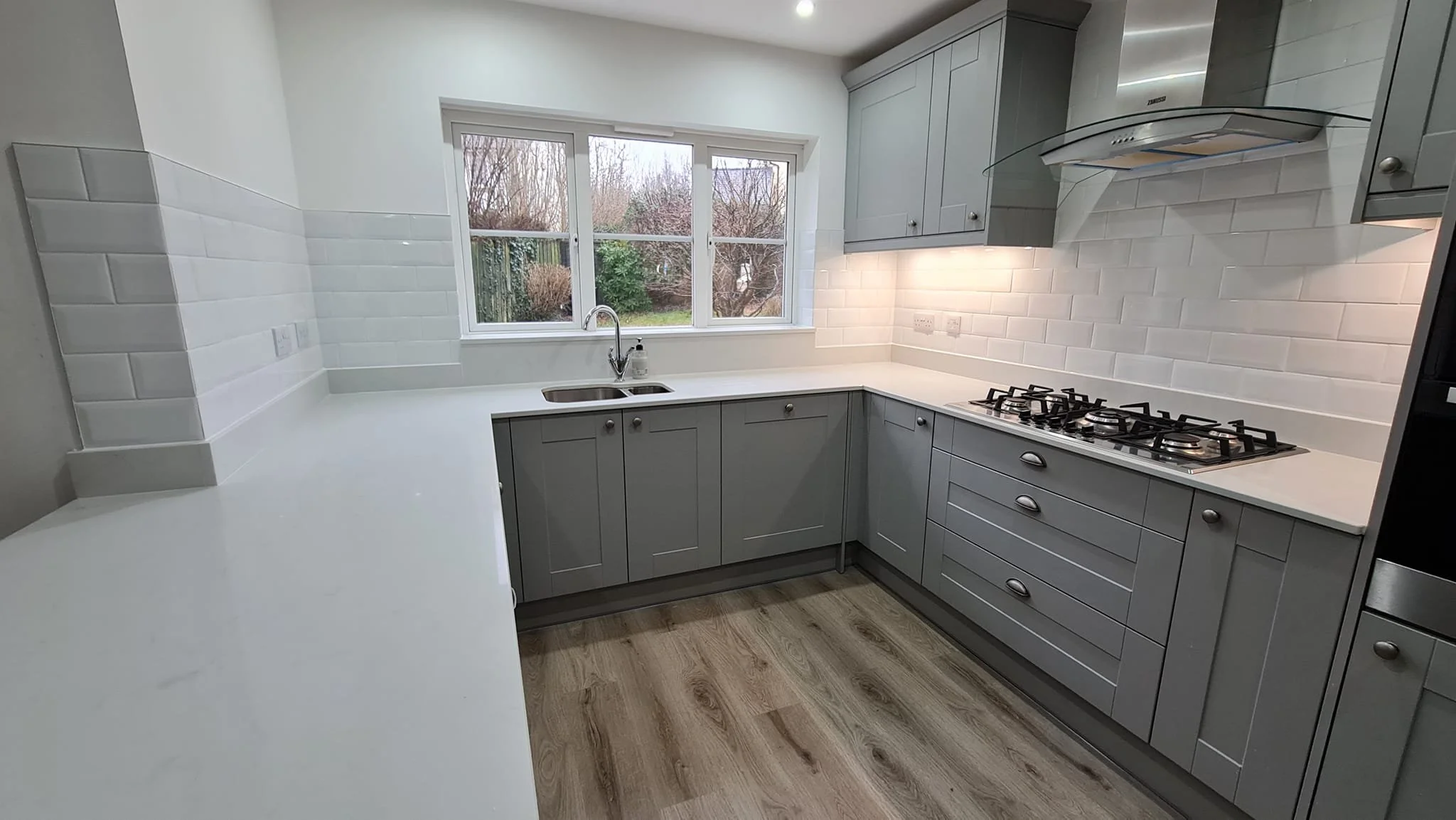 Modern kitchen with gray cabinets, white countertop, window overlooking outdoor garden, stainless steel stove, and wooden flooring.