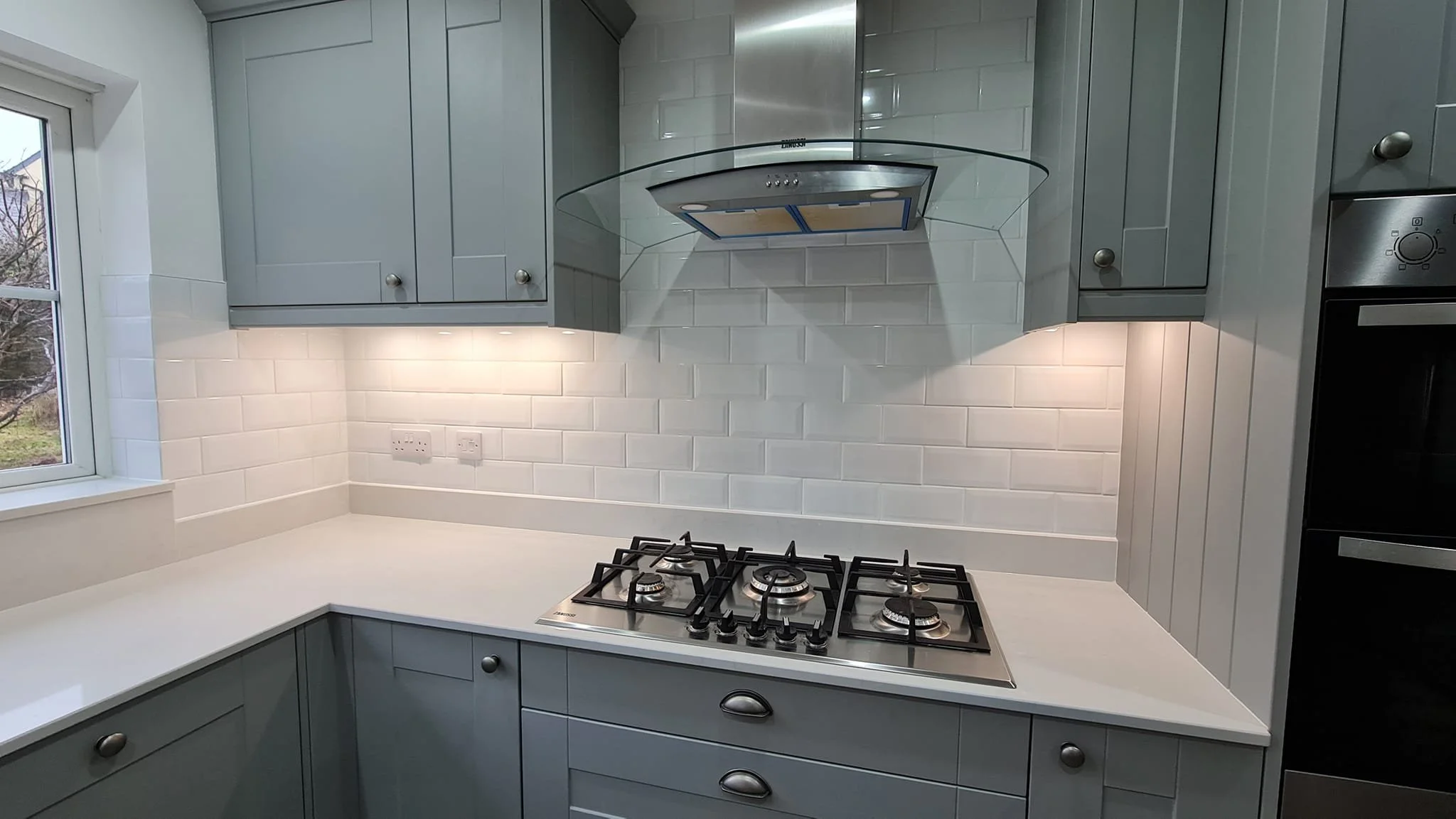 A modern kitchen with gray cabinets, white subway tile backsplash, a gas stove with four burners, and a stainless steel range hood. There is a window on the left side and electrical outlets on the backsplash.