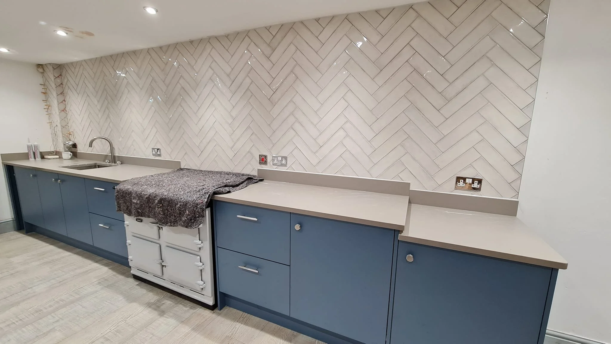 A kitchen with blue lower cabinets, a beige countertop, a white stove with a towel draped over it, and a herringbone-patterned tile backsplash in shades of beige and white.