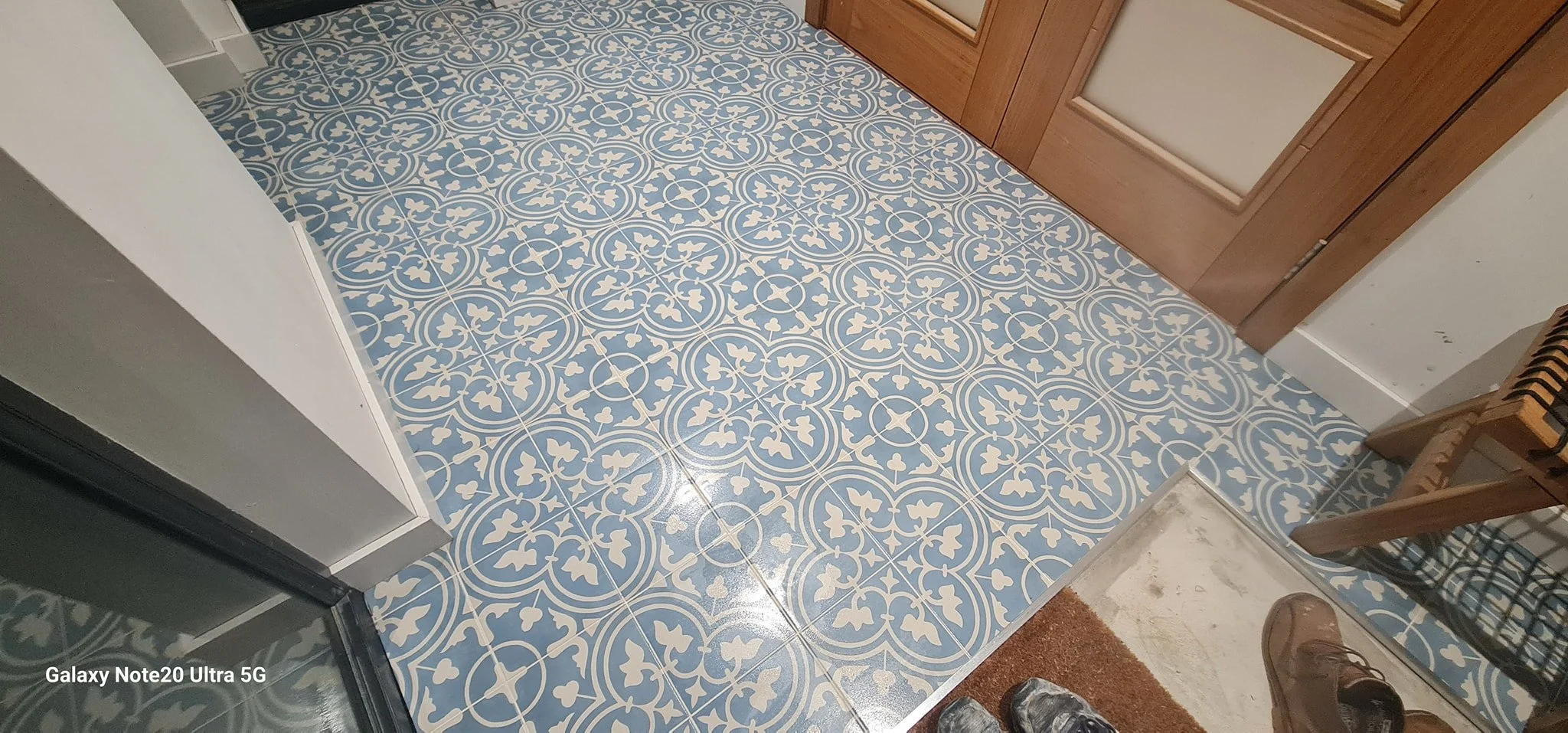 View of patterned blue and white tile flooring next to a wooden door and a shoe rack, with shoes and a person’s feet visible.