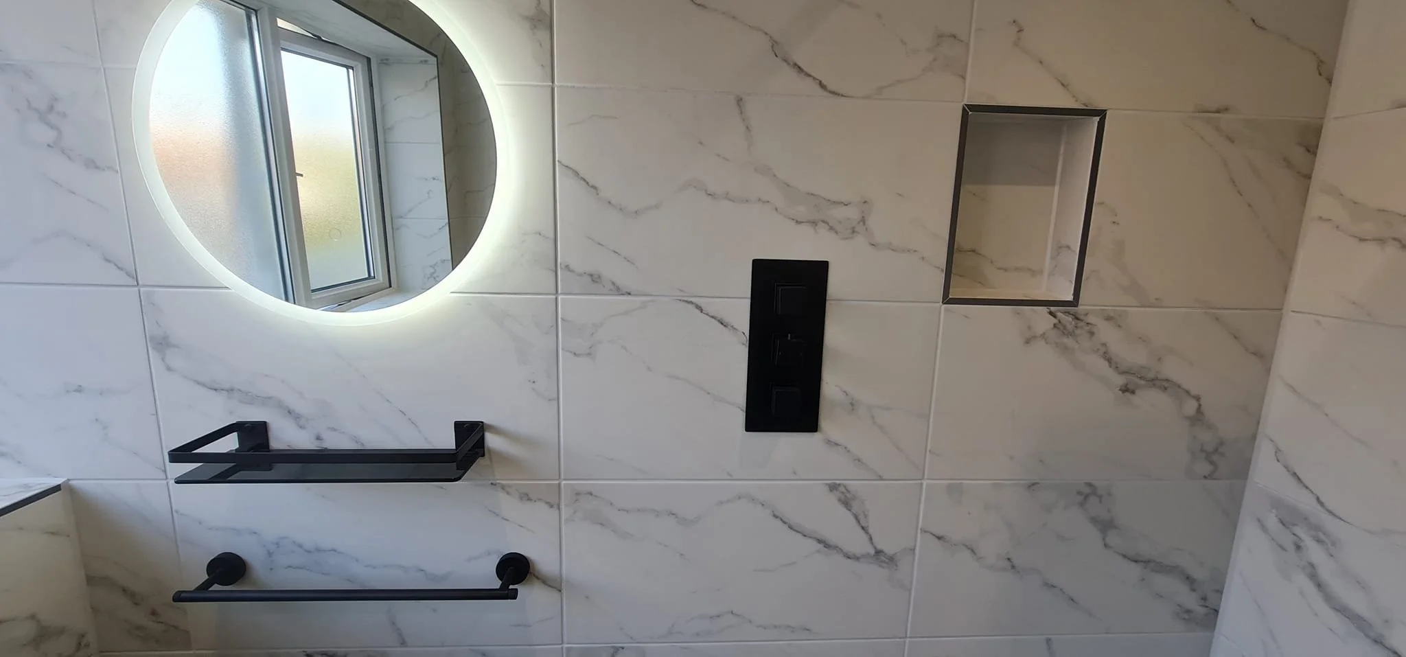 Bathroom wall with large marble tiles, a frosted window, a round backlit mirror, a black towel rack, a black triple switch, and a recessed niche in the wall.
