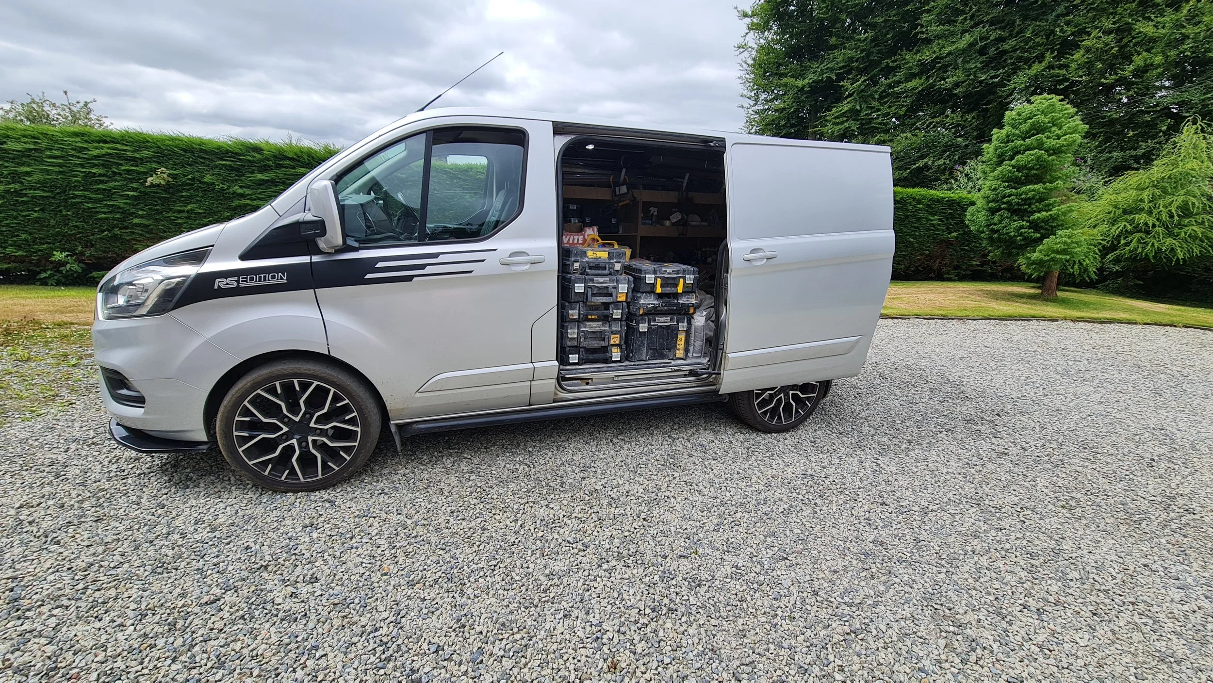 A silver van with its side door open, revealing tools and equipment inside, parked on a gravel driveway with green hedges and trees in the background.