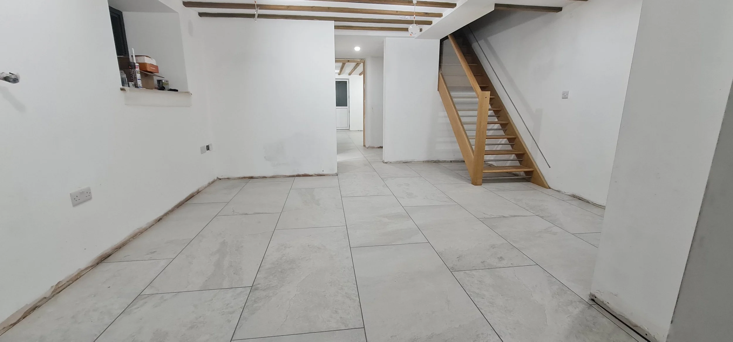 Empty room with white walls, tiled floor, a staircase with wooden steps, and some construction materials on a small windowsill.