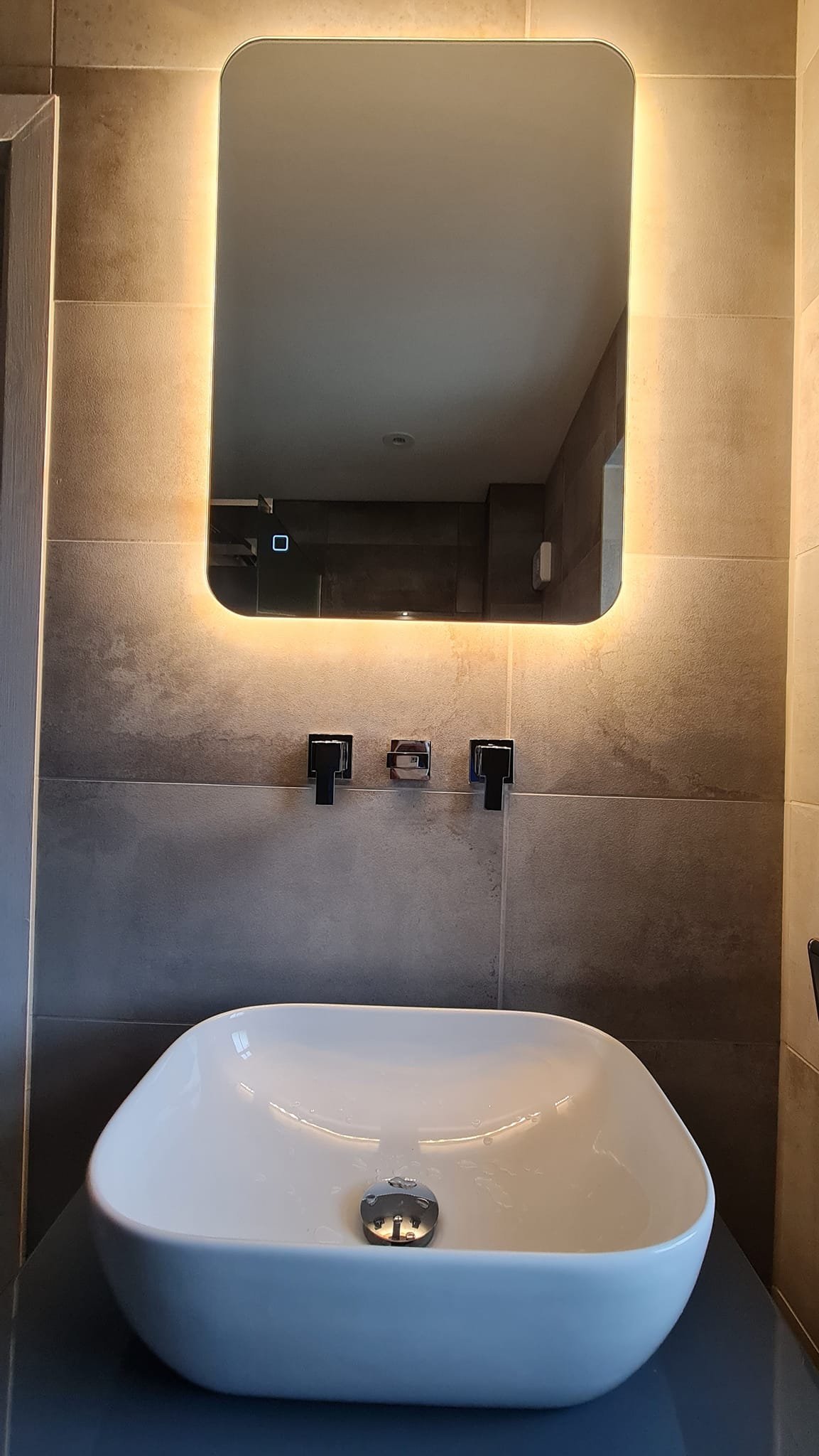 Modern bathroom with a white oval-shaped vessel sink, a rectangular mirror with backlit lighting, and black fixtures against beige tiled walls.